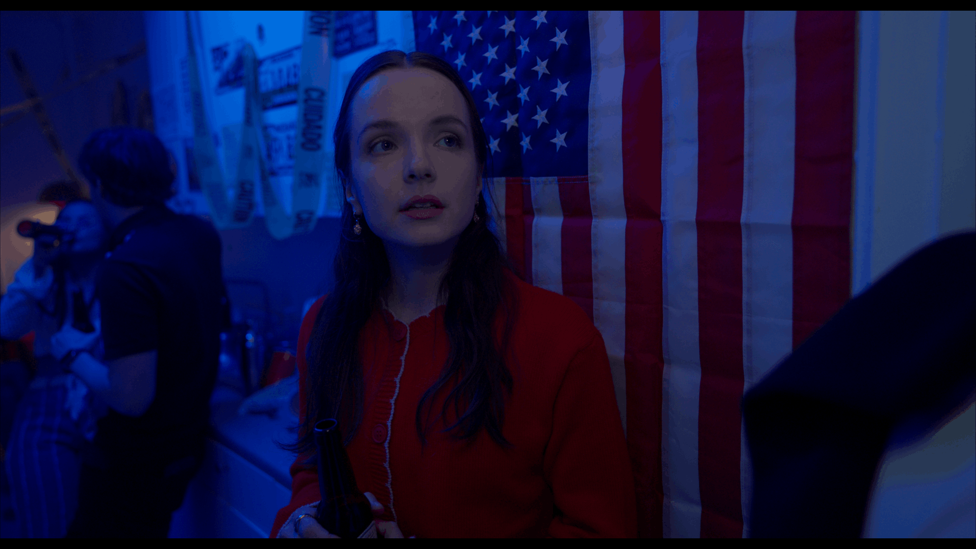A young woman with long dark hair, standing next to an American flag, holds a beer bottle and looks to the side. She is in a dimly lit room with a blue hue, with a man in the background wearing a dark shirt and striped pants.