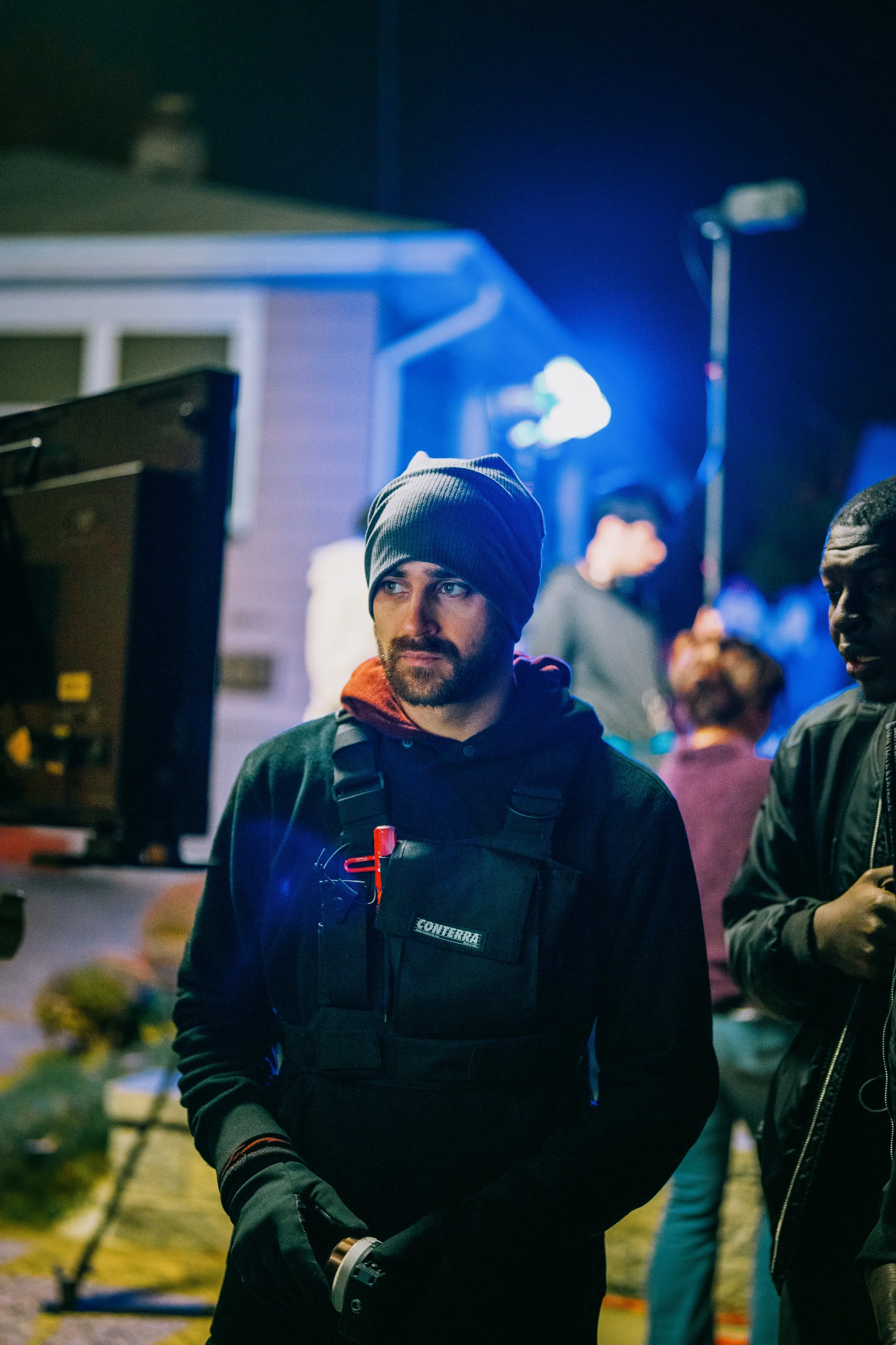 A man wearing a gray beanie, black hoodie, and black vest with a red clip and phone in a case, stands outside at night near a monitor racking focus, with people and equipment around him.