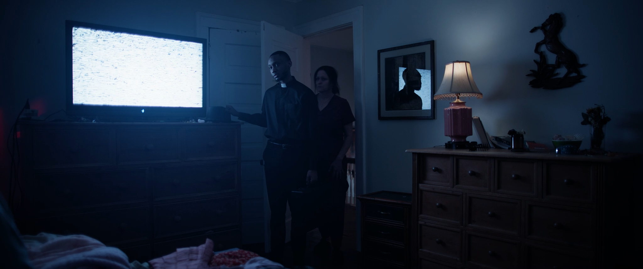 A dark room with a television showing static, a dresser, a lamp on a sideboard, and two people standing near a doorway. One person is dressed as a priest, and the other is a woman, both looking towards the TV.