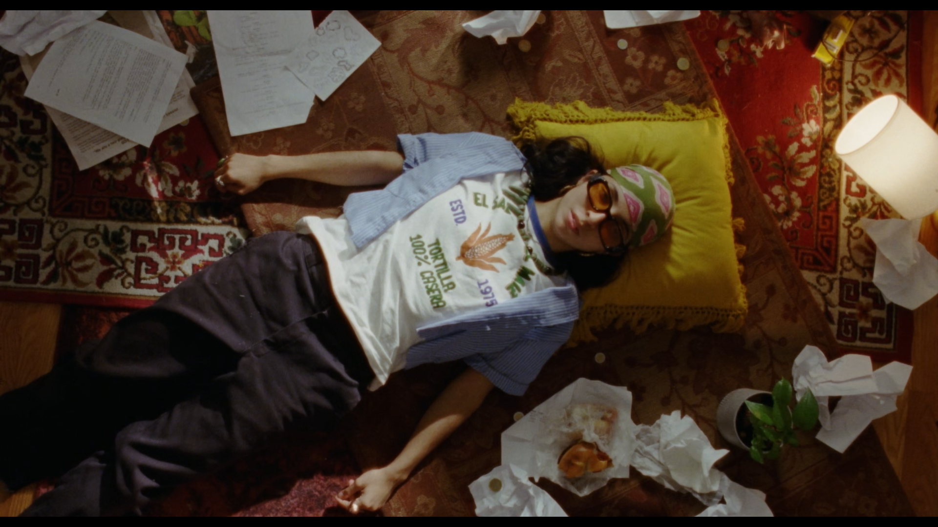 A person lying on their back on a patterned rug surrounded by scattered papers, tissues, a small plant, in a cluttered room, illuminated by a nearby lamp, wearing glasses, a colorful bandana, an oversized T-shirt, and dark pants.