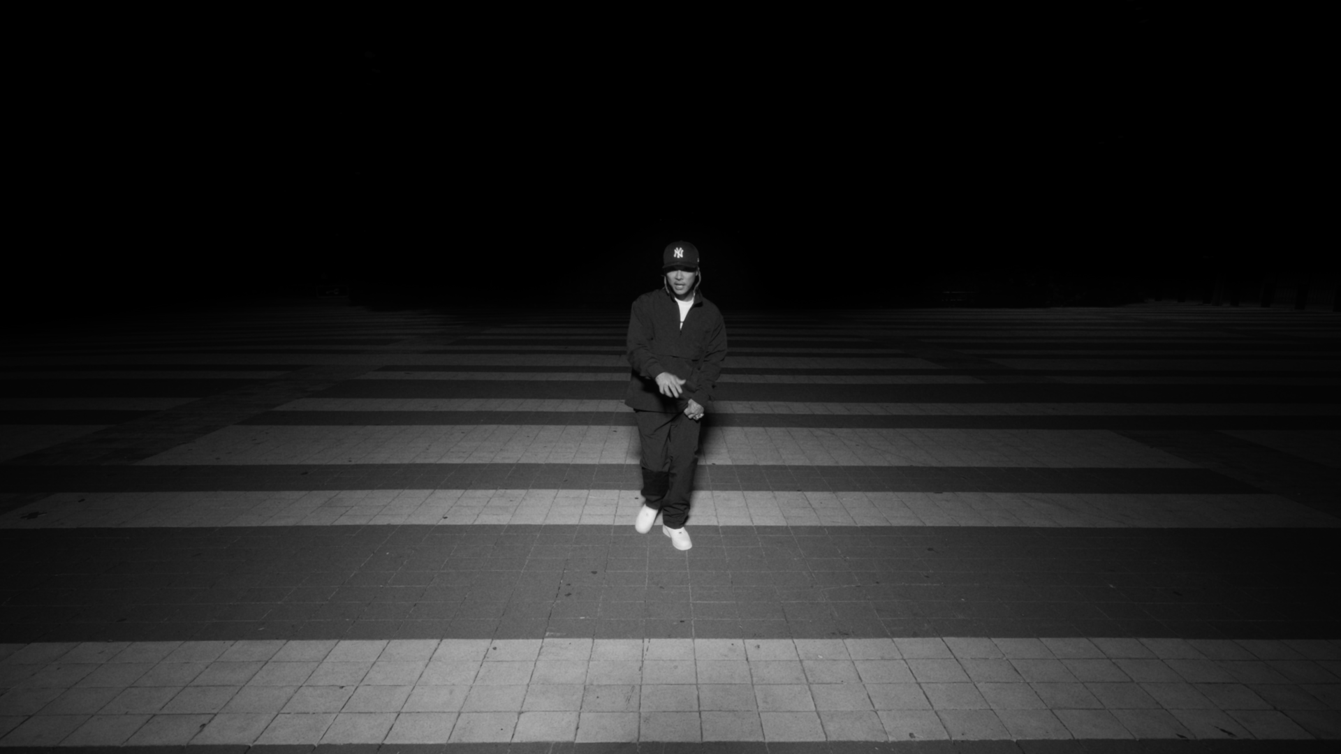 A person standing alone on a crosswalk at night, facing the camera, with darkness surrounding him.