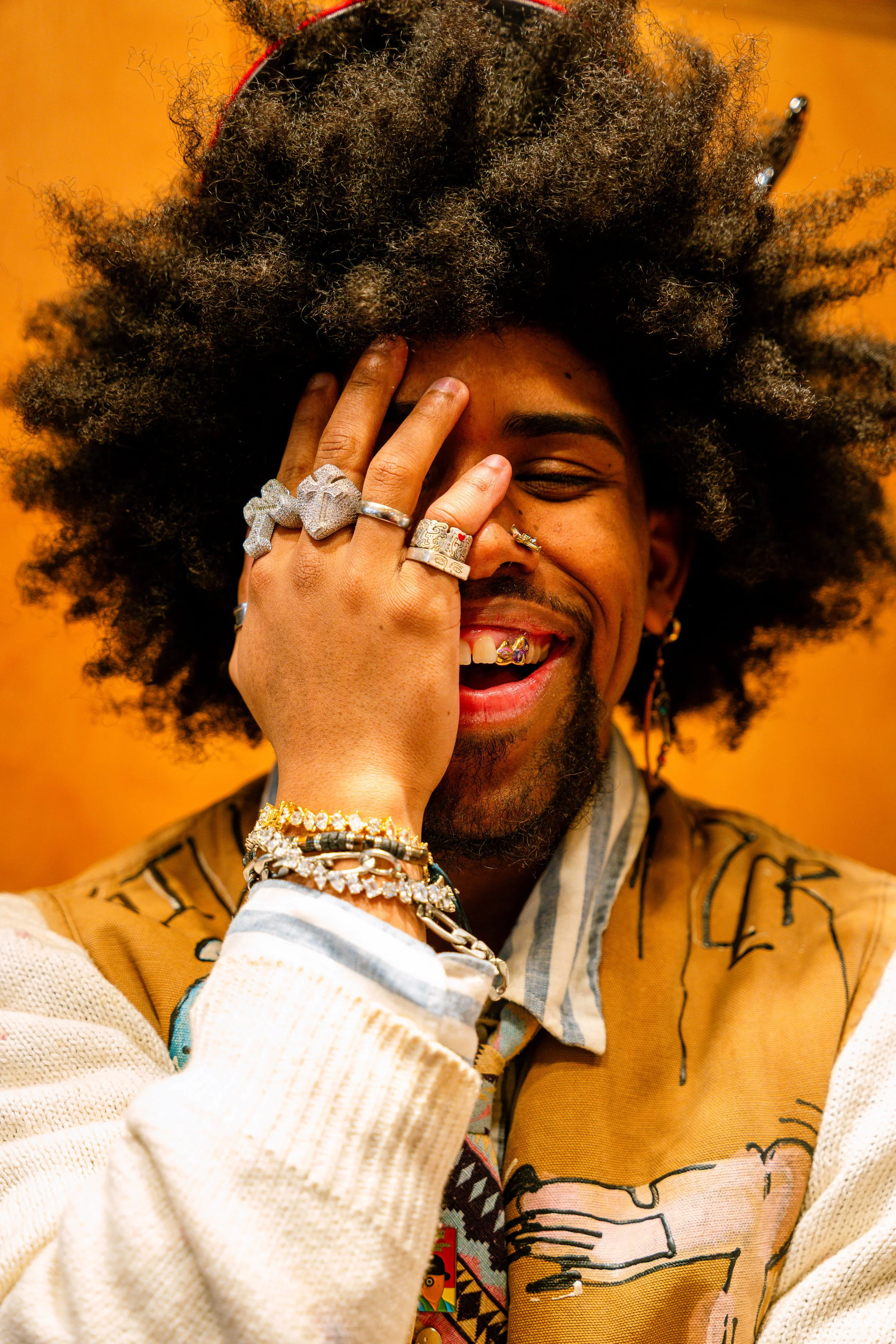 A young man with a large curly afro hairstyle, smiling and covering part of his face with his hand, wearing multiple rings and bracelets, earrings, and a colorful, patterned shirt.