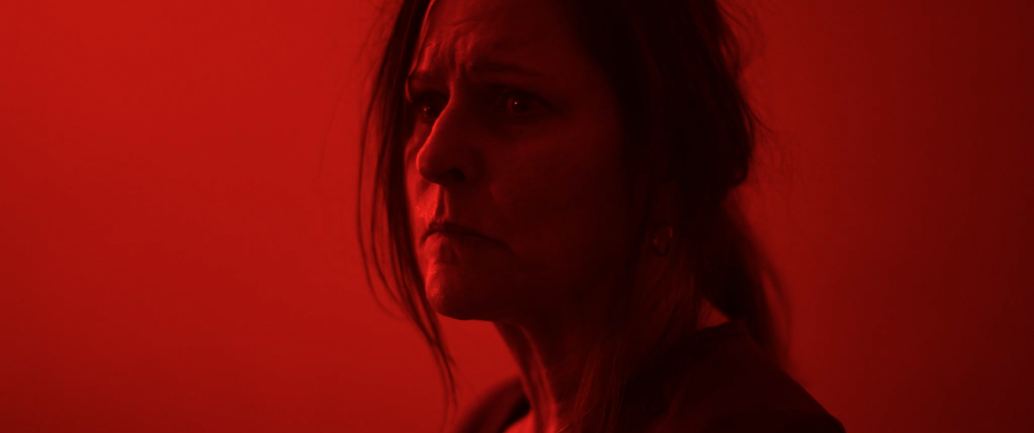 A woman with long hair and earrings, viewed in profile against a red background, looking serious or contemplative.