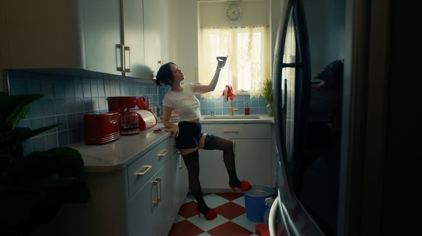 A woman leaning against a kitchen counter, dressed in black stockings, red high heels, a white t-shirt, and red gloves, holding a glass in a sunlit kitchen with blue tiles and cream cabinets.