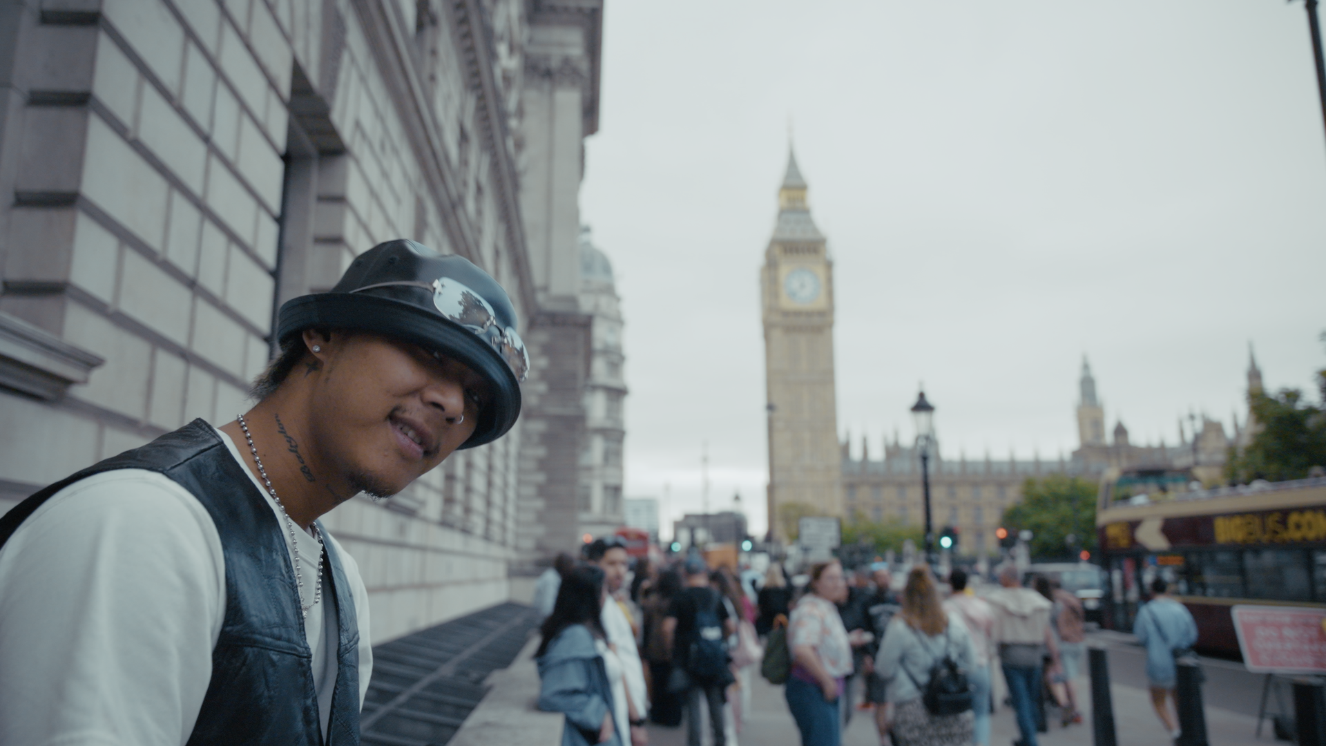 A young man wearing a black leather cap, black sunglasses, and a black leather vest over a white shirt, standing on a busy London street near Big Ben with a crowd of people and a red double-decker bus in the background.