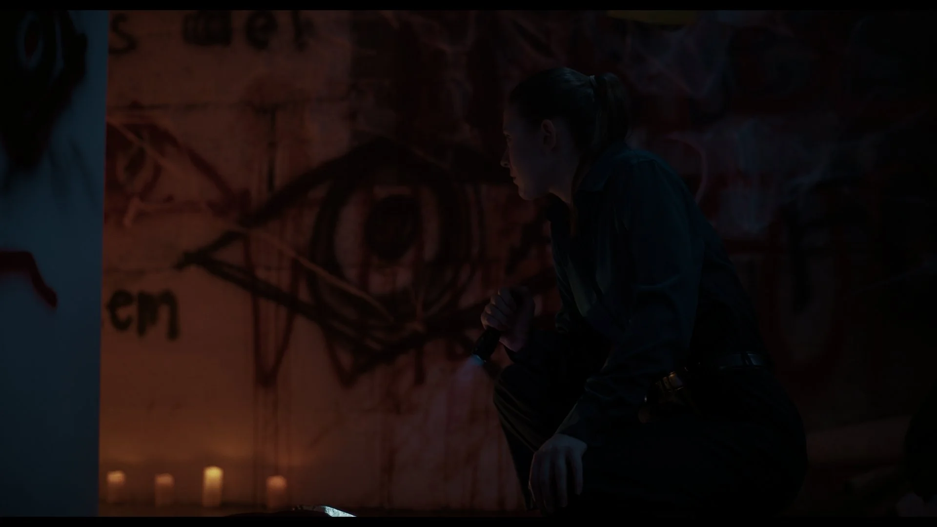 A woman crouching down in a dark room, holding a flashlight, with graffiti on the wall behind her.