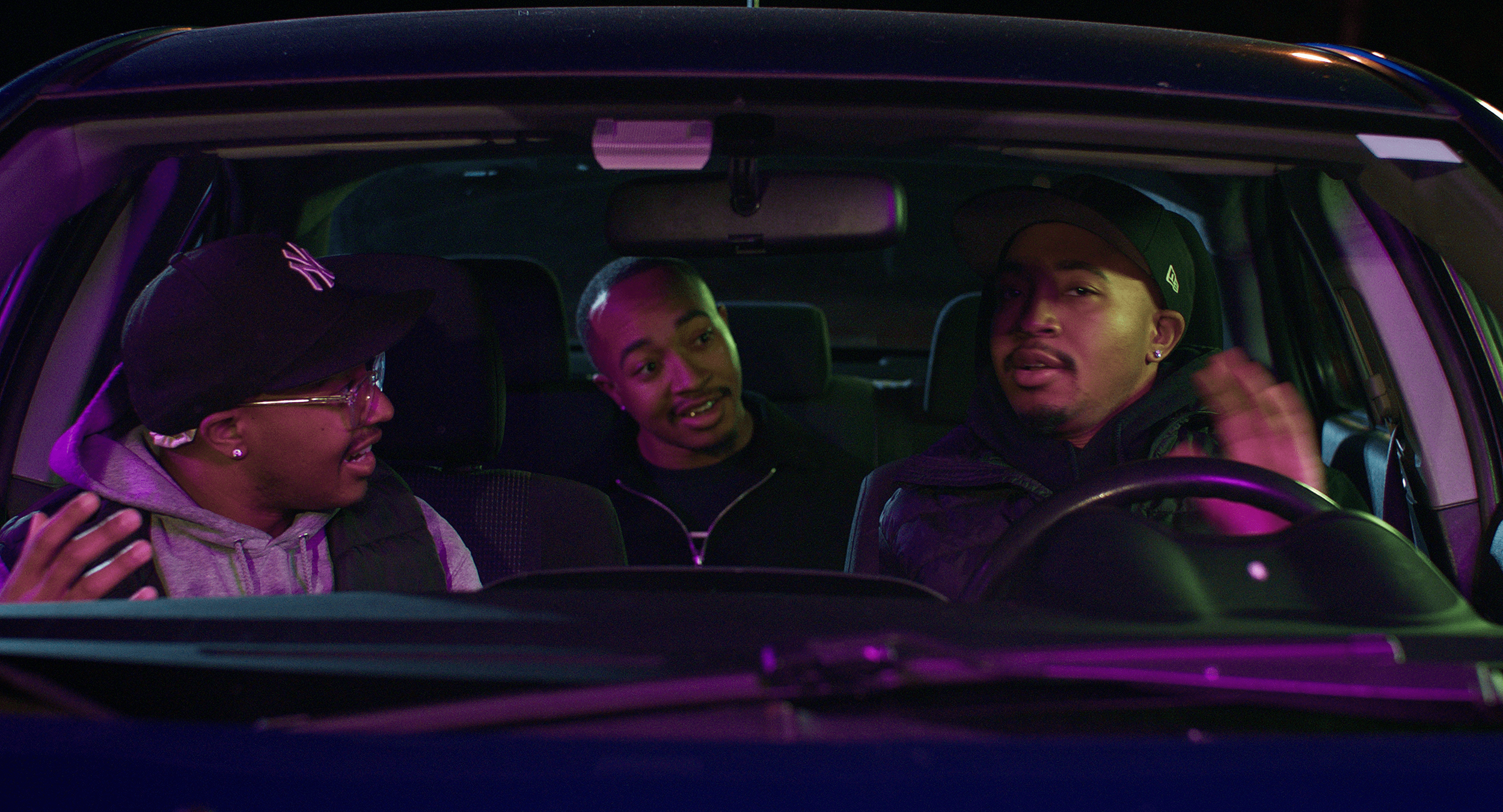 Three young men inside a car at night, engaging in conversation, with colorful purple and green lighting.