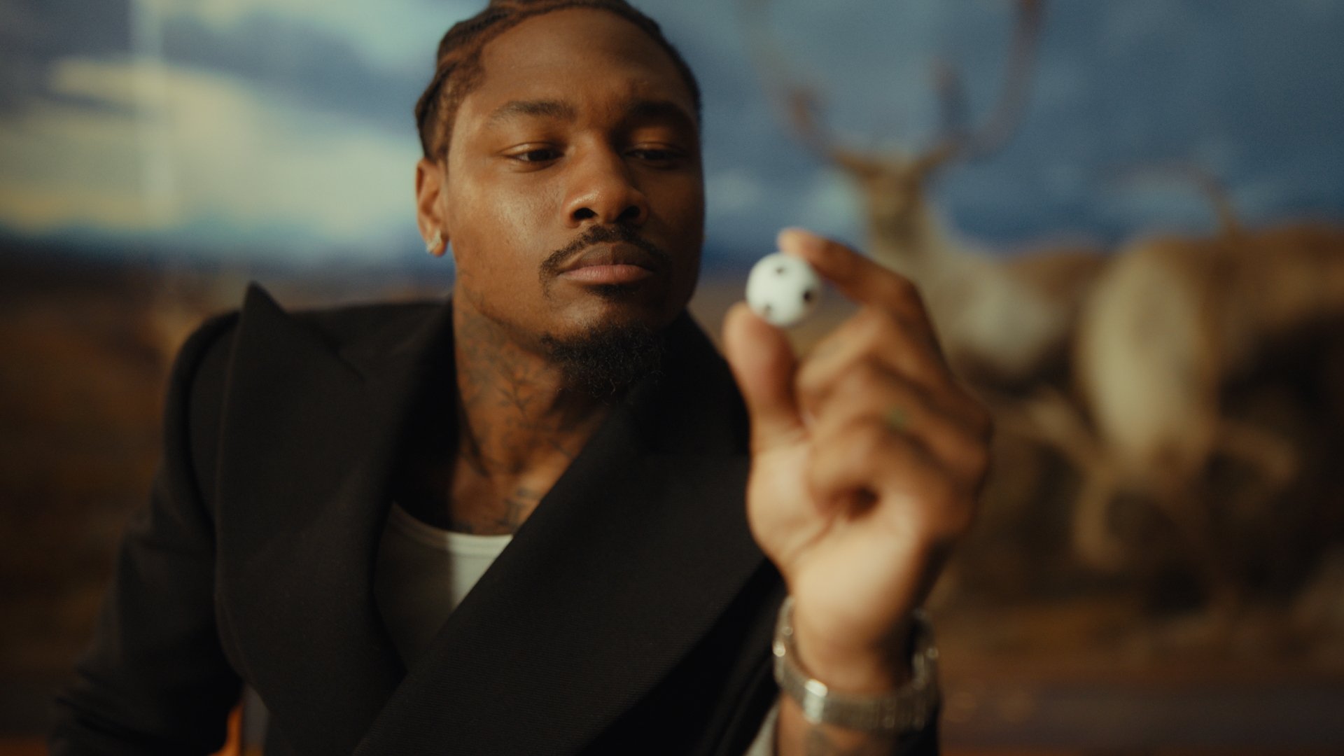 A man with tattoos, wearing a black blazer and a white shirt, looking at a small white skull in his hand, with a blurred background of a painting or mural.