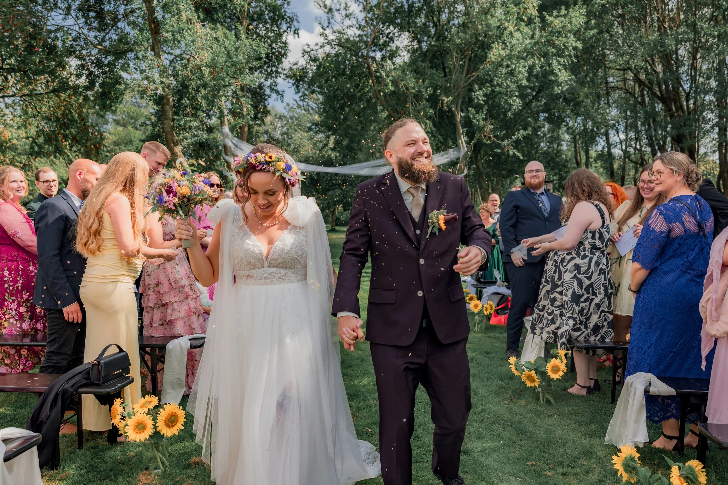 Et bryllupsceremoni med brudepar, der går hånd i hånd, omgivet af venner og familie i en grøn park. Bruden holder en blomsterbuket, og gæsterne smiler og kaster konfetti.