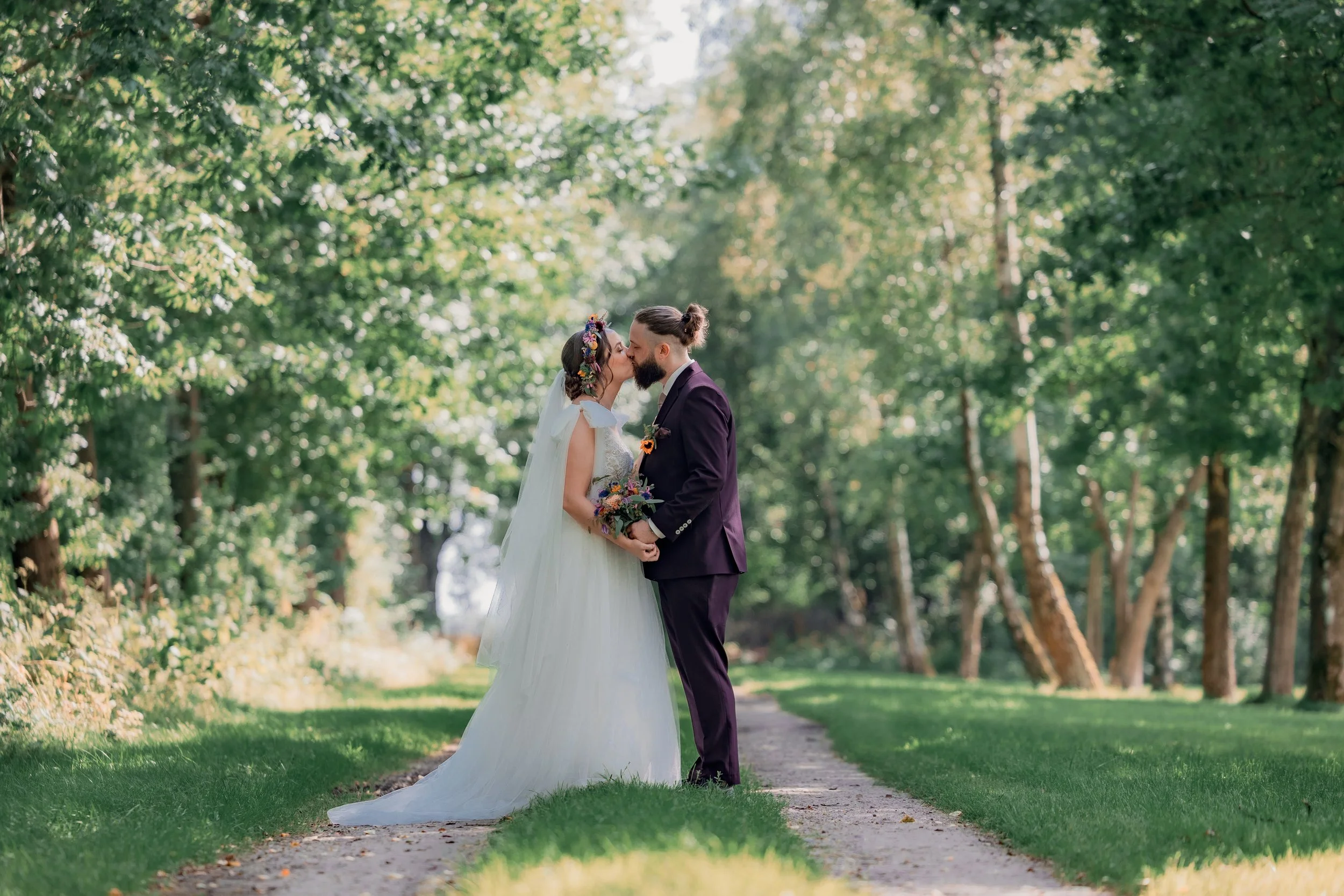 Et brudepar står på en sti i en grøn skov, de kysser hinanden og holder i hænder. Bruden bærer en hvid kjole med slør og har en blomsterkrans i håret, mens brudgommen har en mørkeblå dragt.