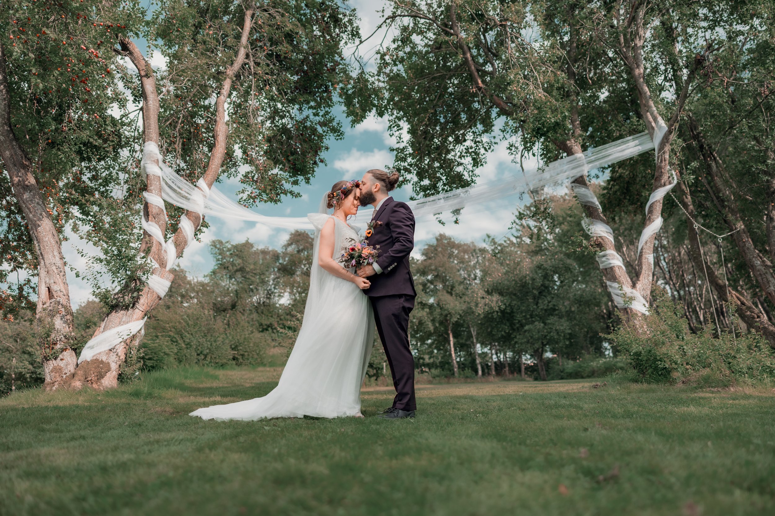 Et bryllupsceremoni udendørs med et par, der står tæt sammen under træer med hvide bånd, på en grøn eng, med blå himmel og skyer i baggrunden.