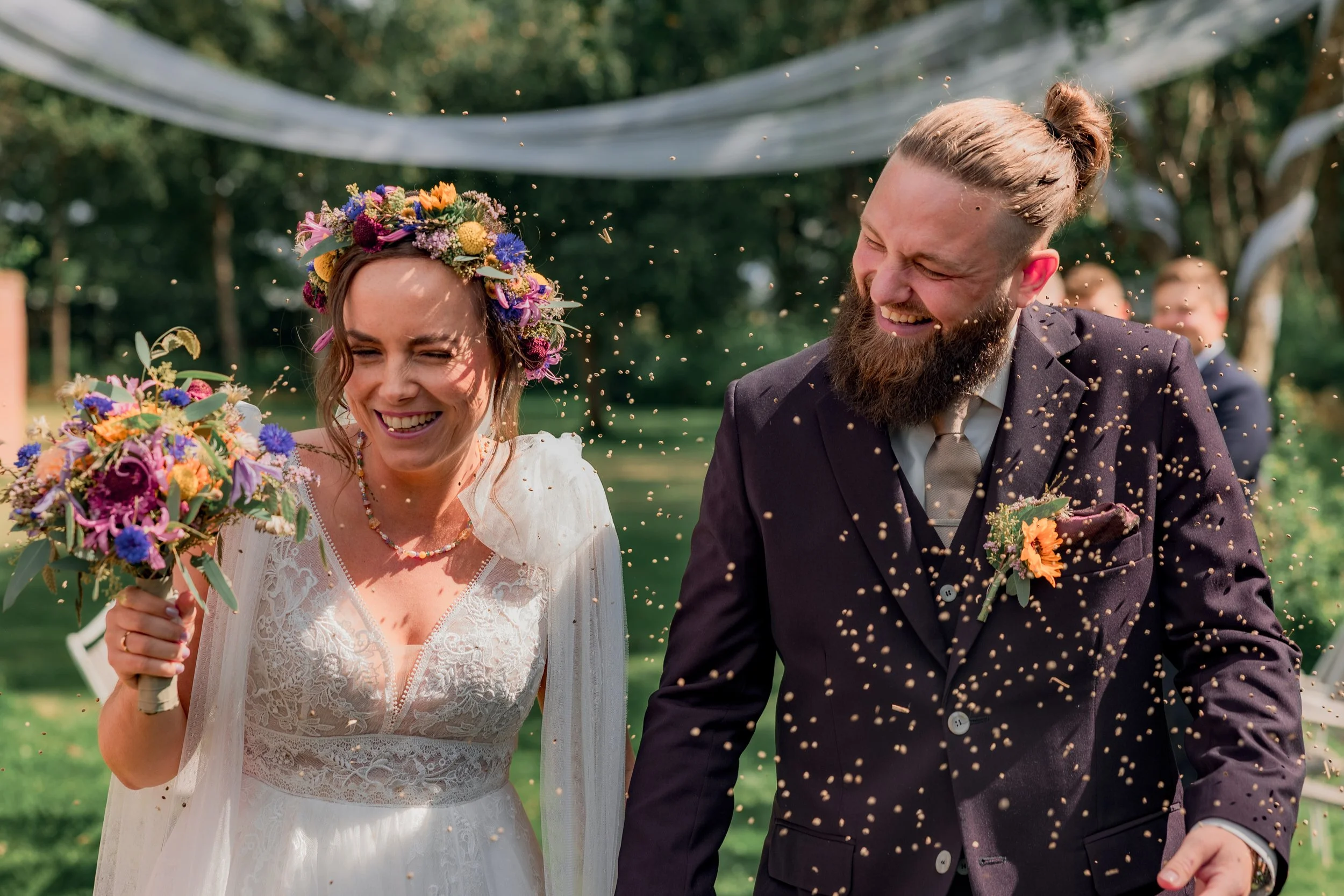 En brud og en brudgom smiler og griner under en bryllupsfest udendørs. Bruden bærer en hvid kjole og en blomsterkrans, mens brudgommen har på en mørk jakkesæt med en blomst i lommen. De holder hinanden i hånden, og der er konfetti i luften.