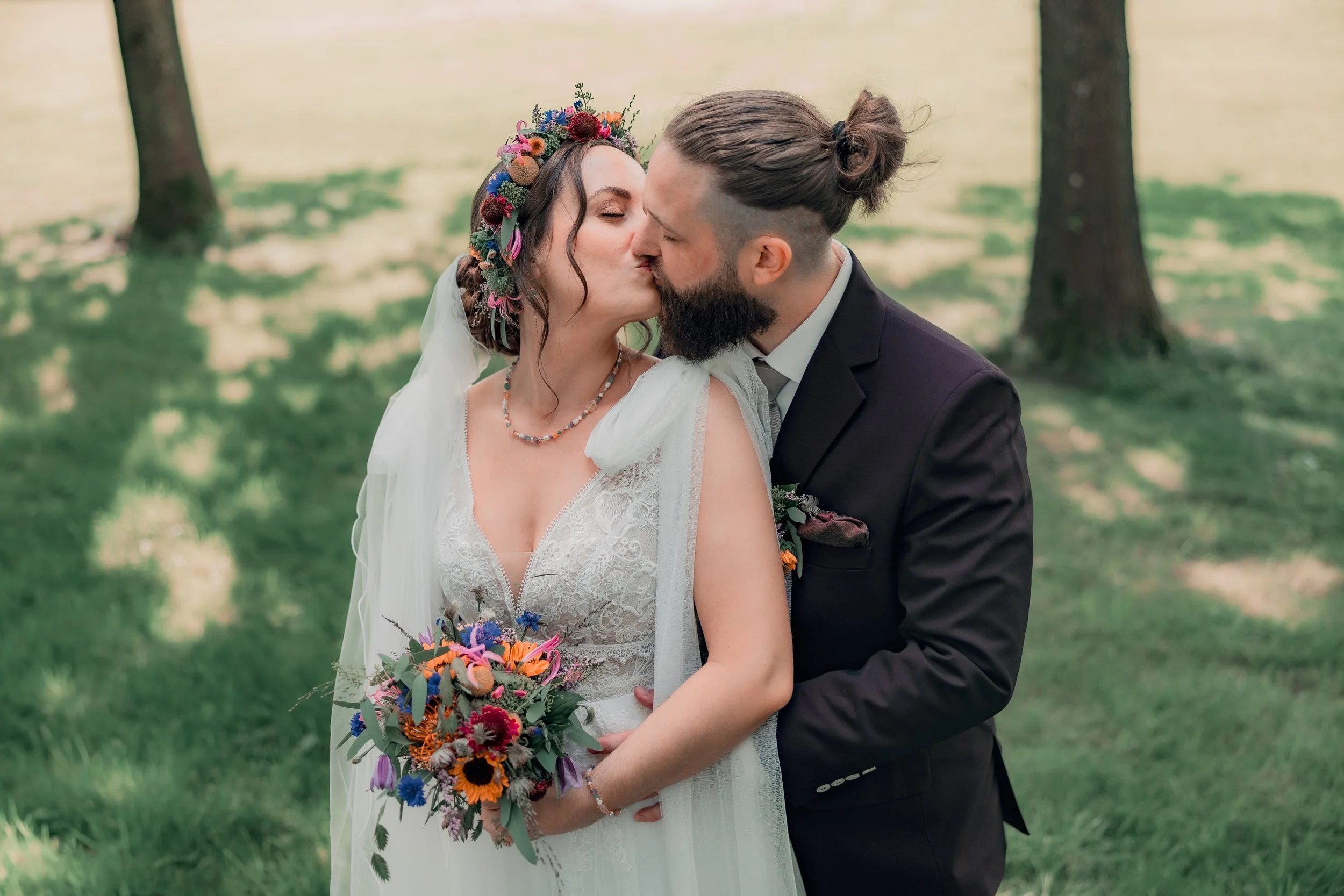Et brudepar kysser hinanden under et træ i en grøn park, med kvinden i en hvid brudekjole med blomsterkrans i håret og manden i en sort jakkesæt.