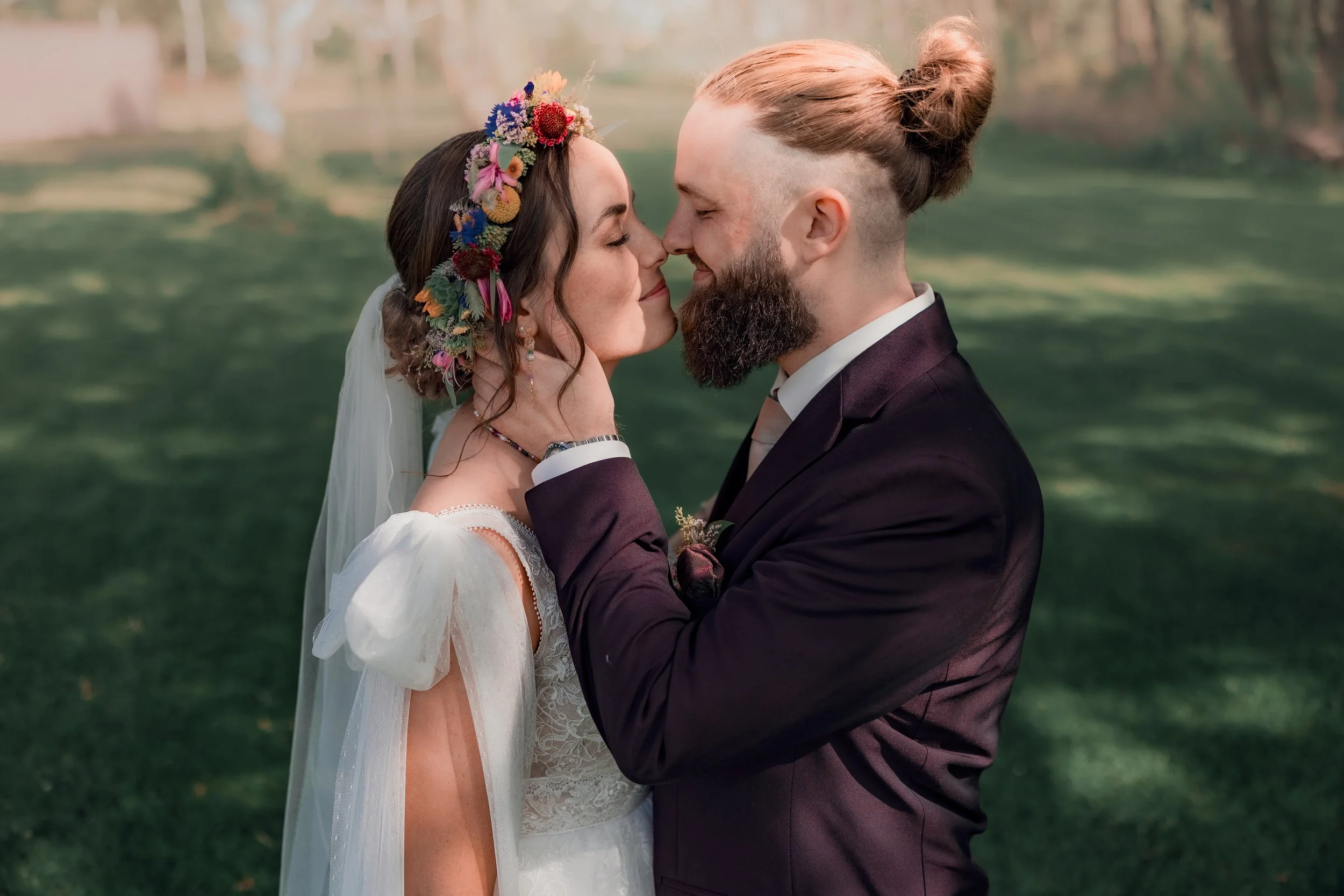 Et brudepar står tæt sammen med ansigterne næsten rørende, i en park med grønne græsplæner og træer i baggrunden.