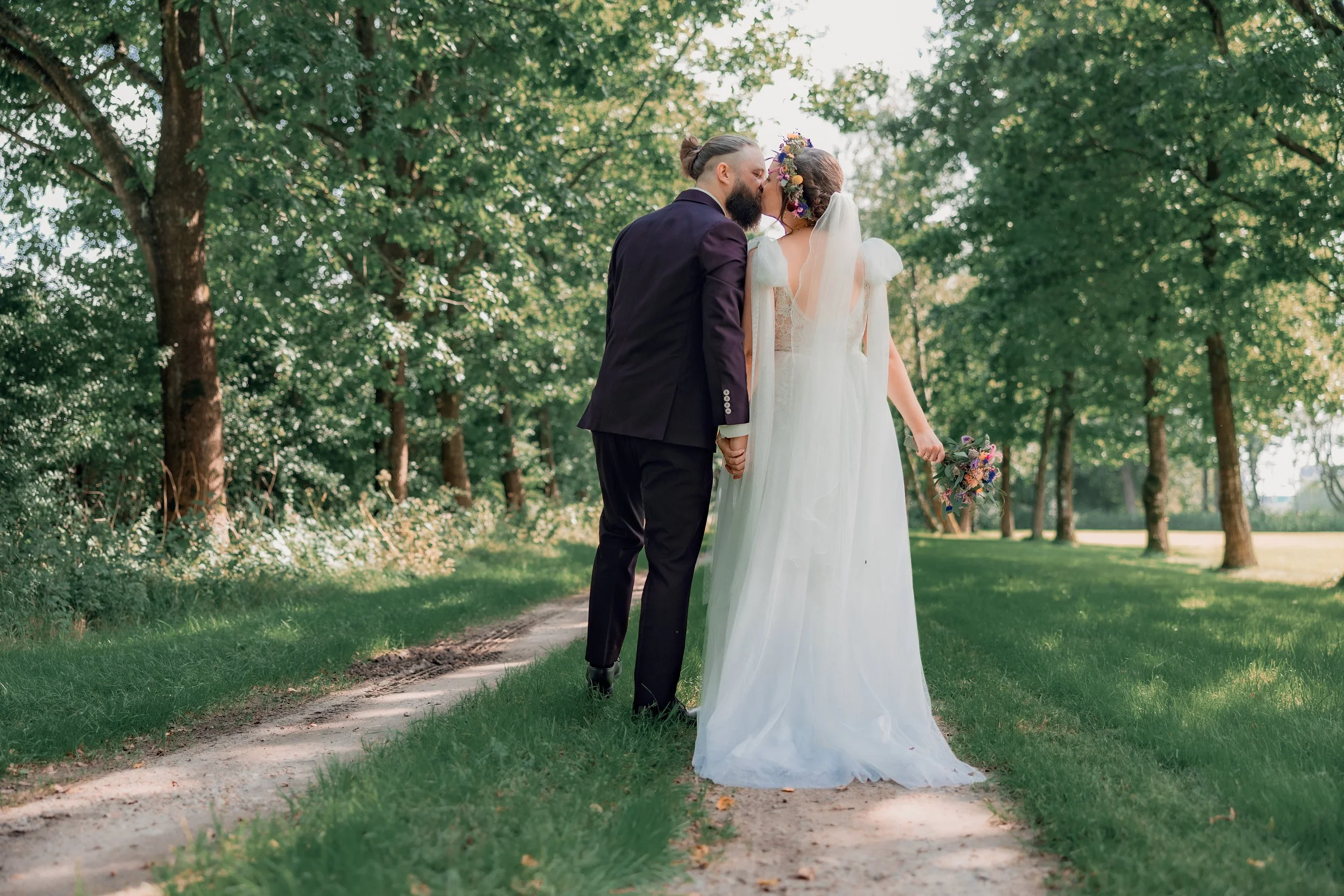 Et brudepar står på en sti i et grønt skovområde, hvor de holder hinanden i hånden og kysser. Kvinden bærer en hvid brudekjole med blomsterkrans i håret og holder en blomsterbuket, mens mannen er i en sort formel habit.
