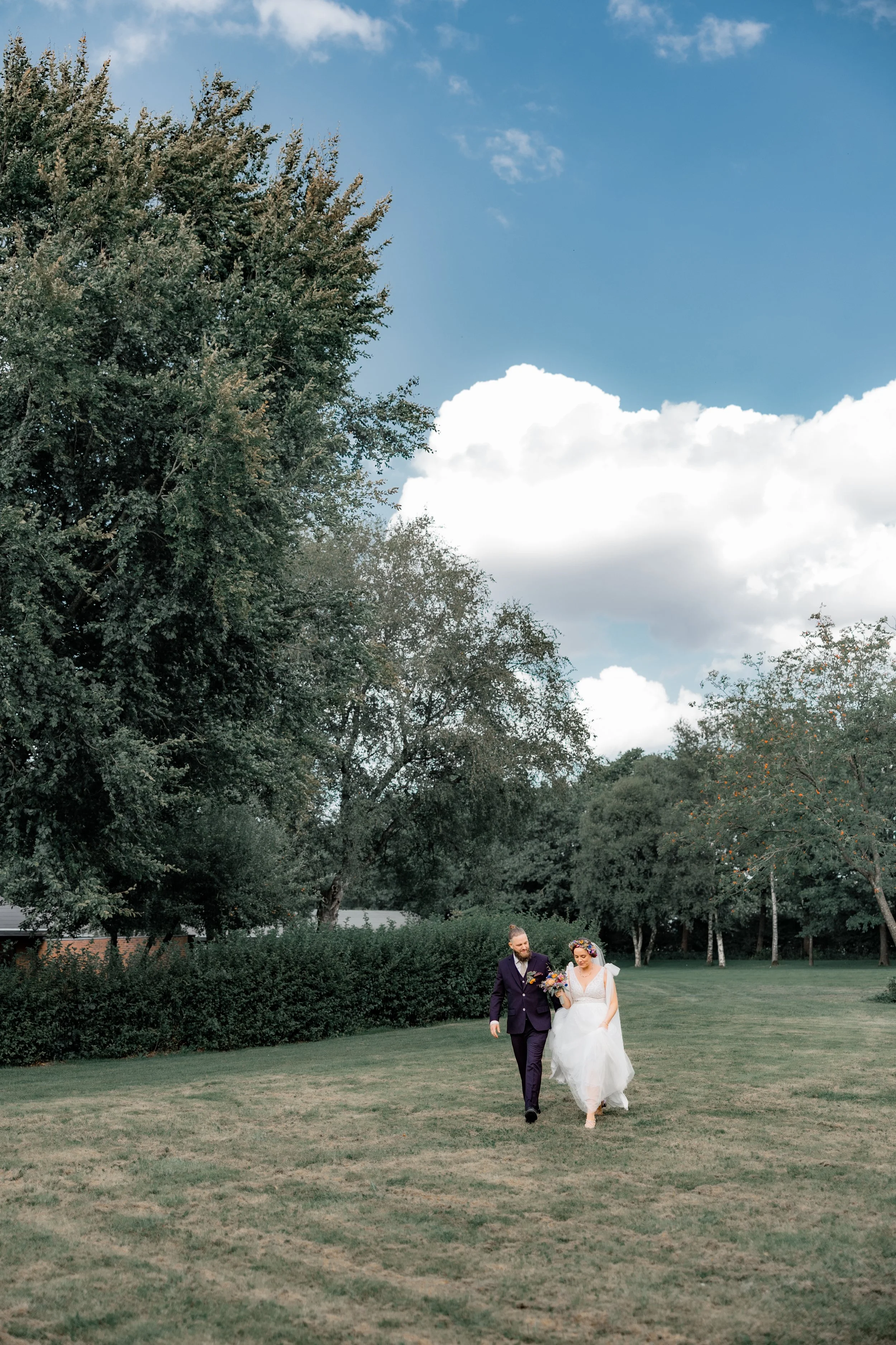 Brudepar går sammen på en grøn parkvej under en blå himmel med spredte skyer, omgivet af store træer og buske.