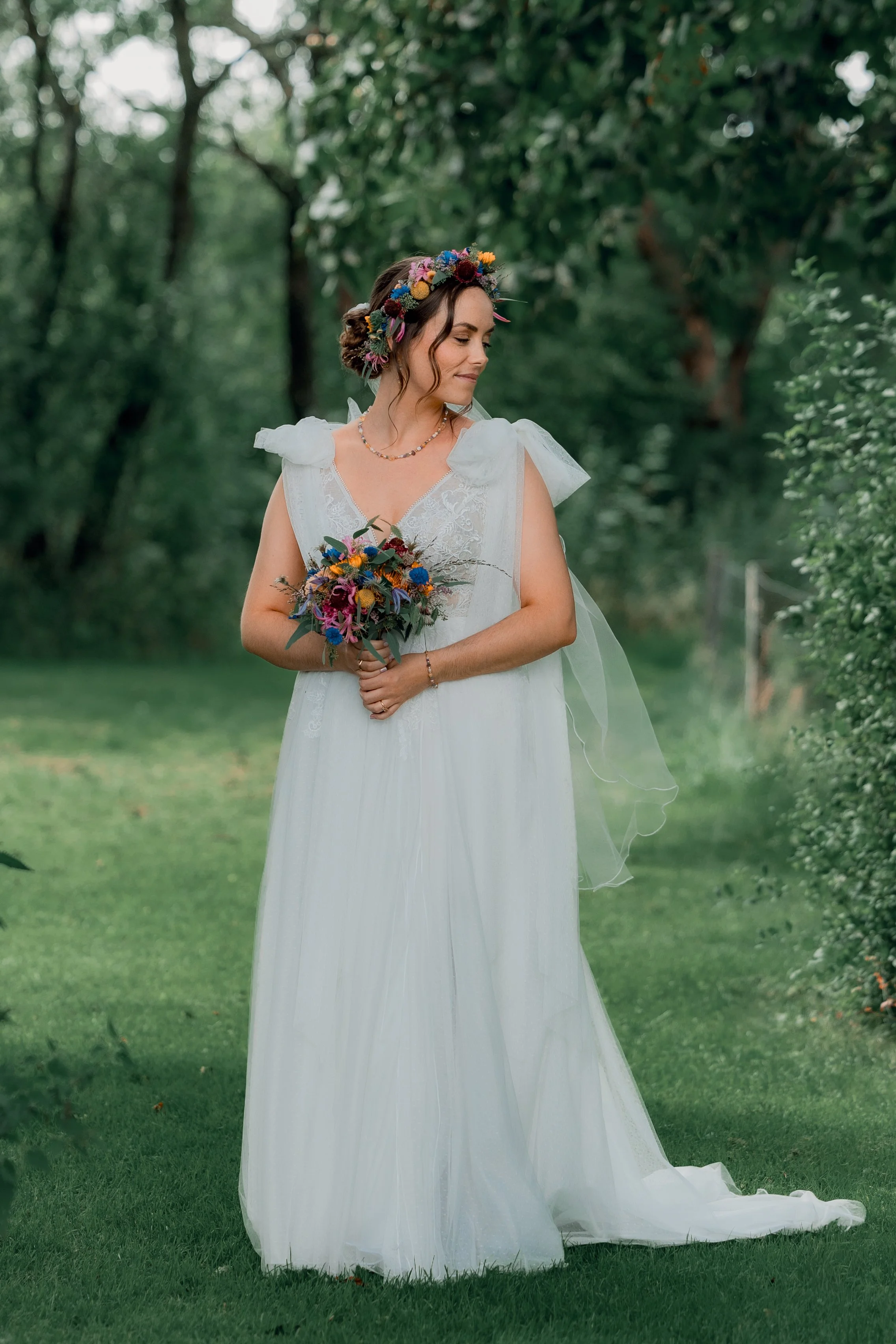 En kvinde i en hvid brudekjole med blomsterkrans i håret holder en blomsterbuket i en grøn skov.
