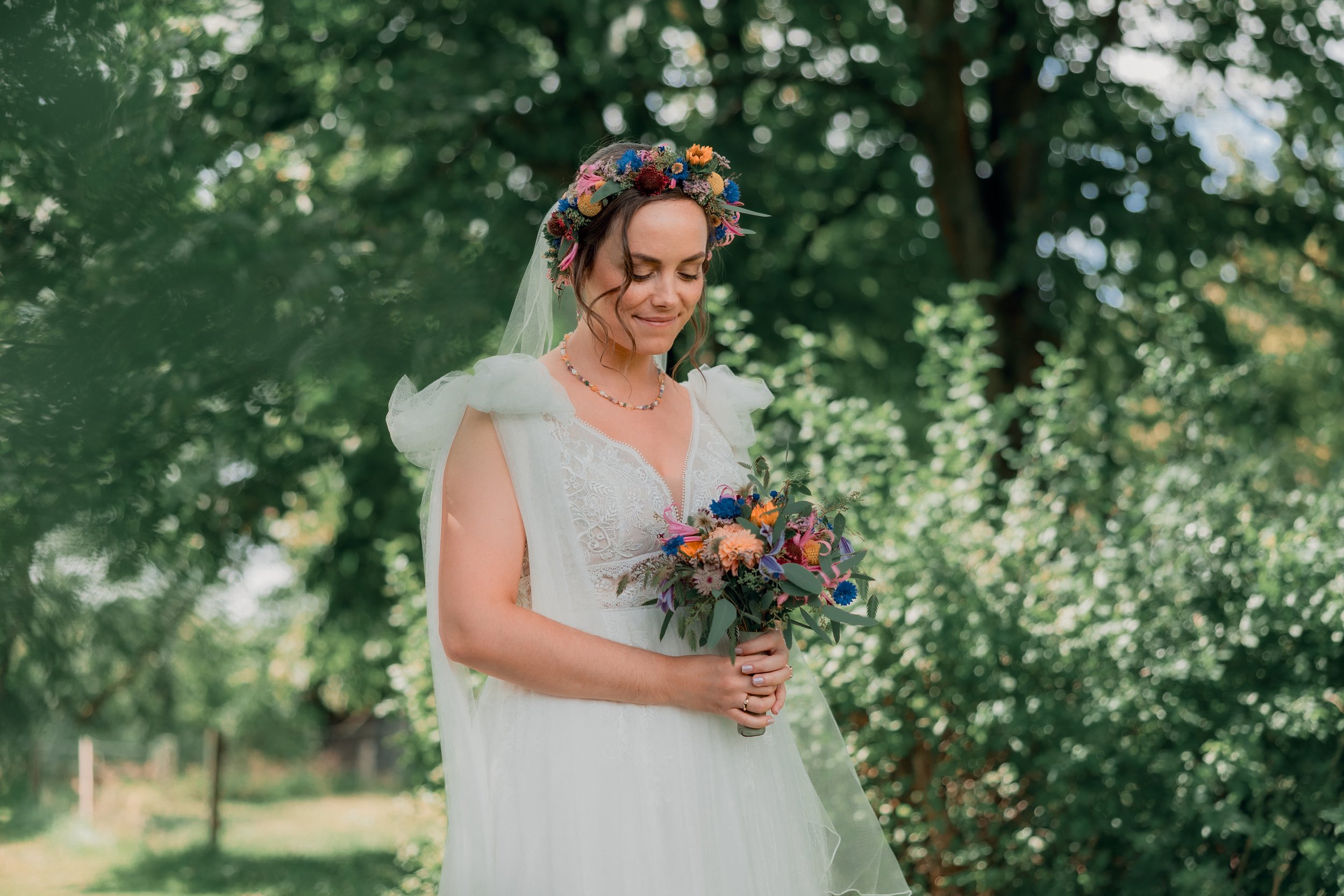 En kvinde i brudekjole foran grønne træer, bærer en blomsterkrans og holder en buket blomster.