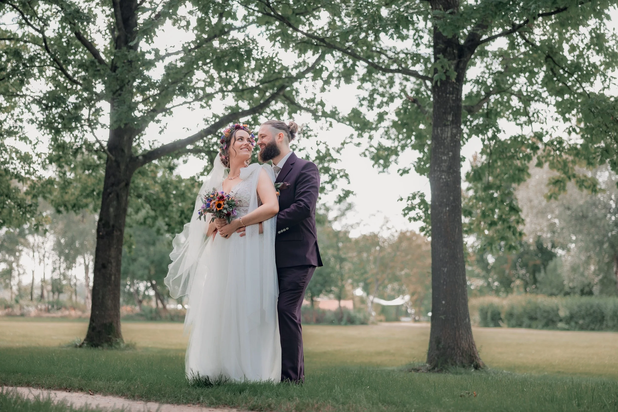 Et brudepar står sammen under store grønne træer i en park, den unge kvinde i hvid brudekjole med blomsterguirlande i håret, den unge mand i mørk habit, de smiler til hinanden, kvinden holder en blomsterbuket.