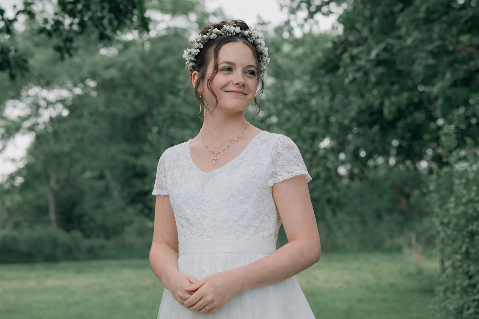 En glad ung kvinde i brudekjole med blomsterkrans på hovedet, står udendørs i en grøn park.