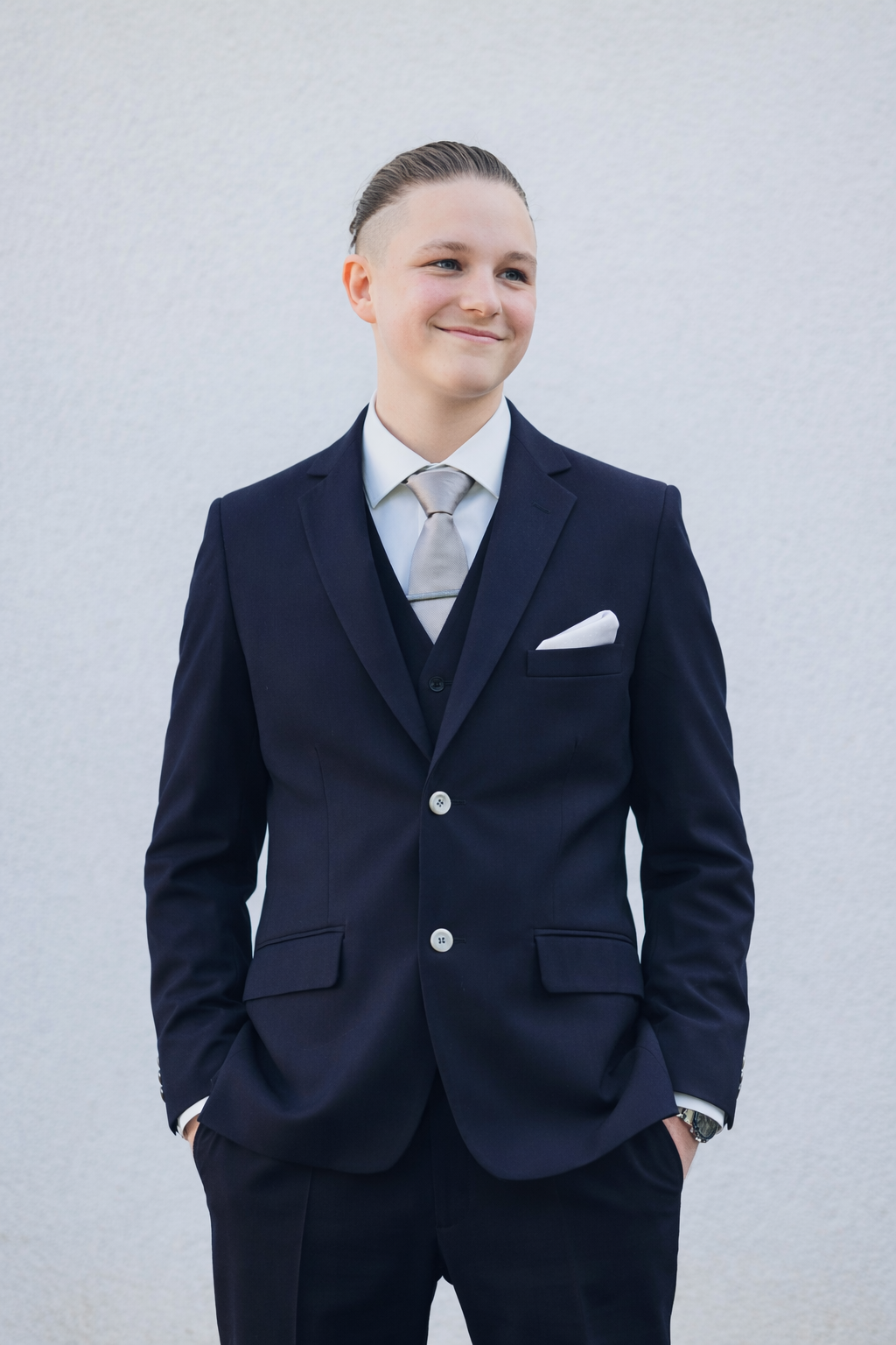 En ung mand i elegant navyblå habit og lysegråt slips står foran en hvid væg, smilende og med hånden i lommen.