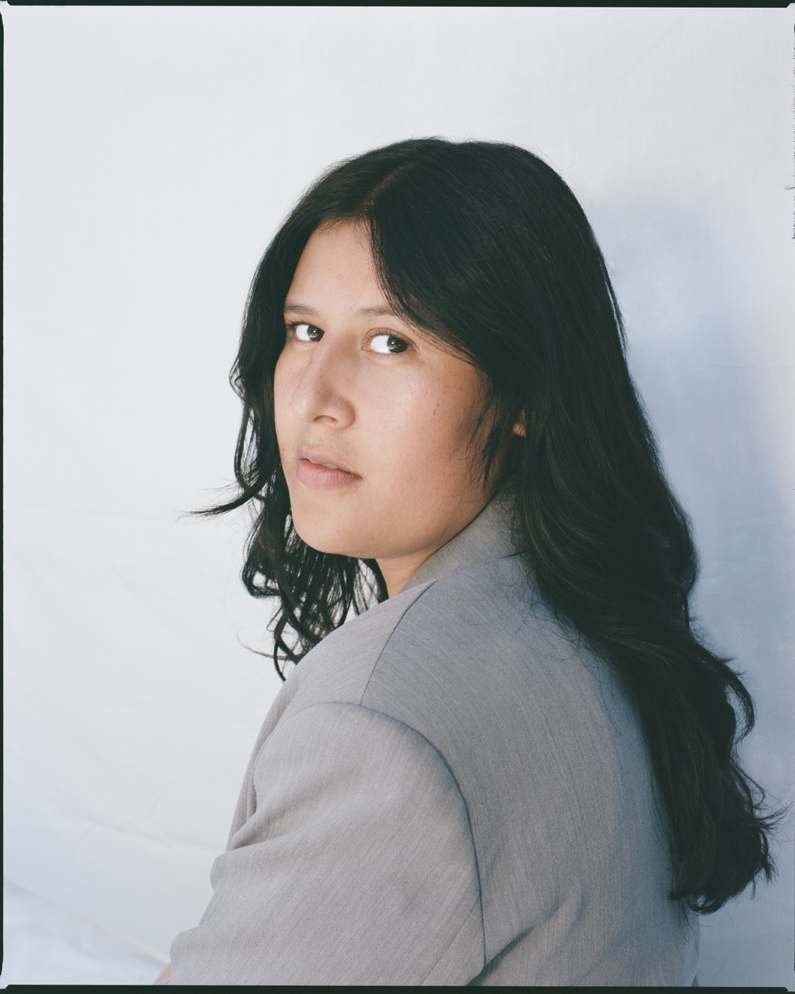 Portrait of a woman with long black wavy hair, wearing a light gray blazer, looking over her shoulder at the camera with a neutral expression, against a plain light background.
