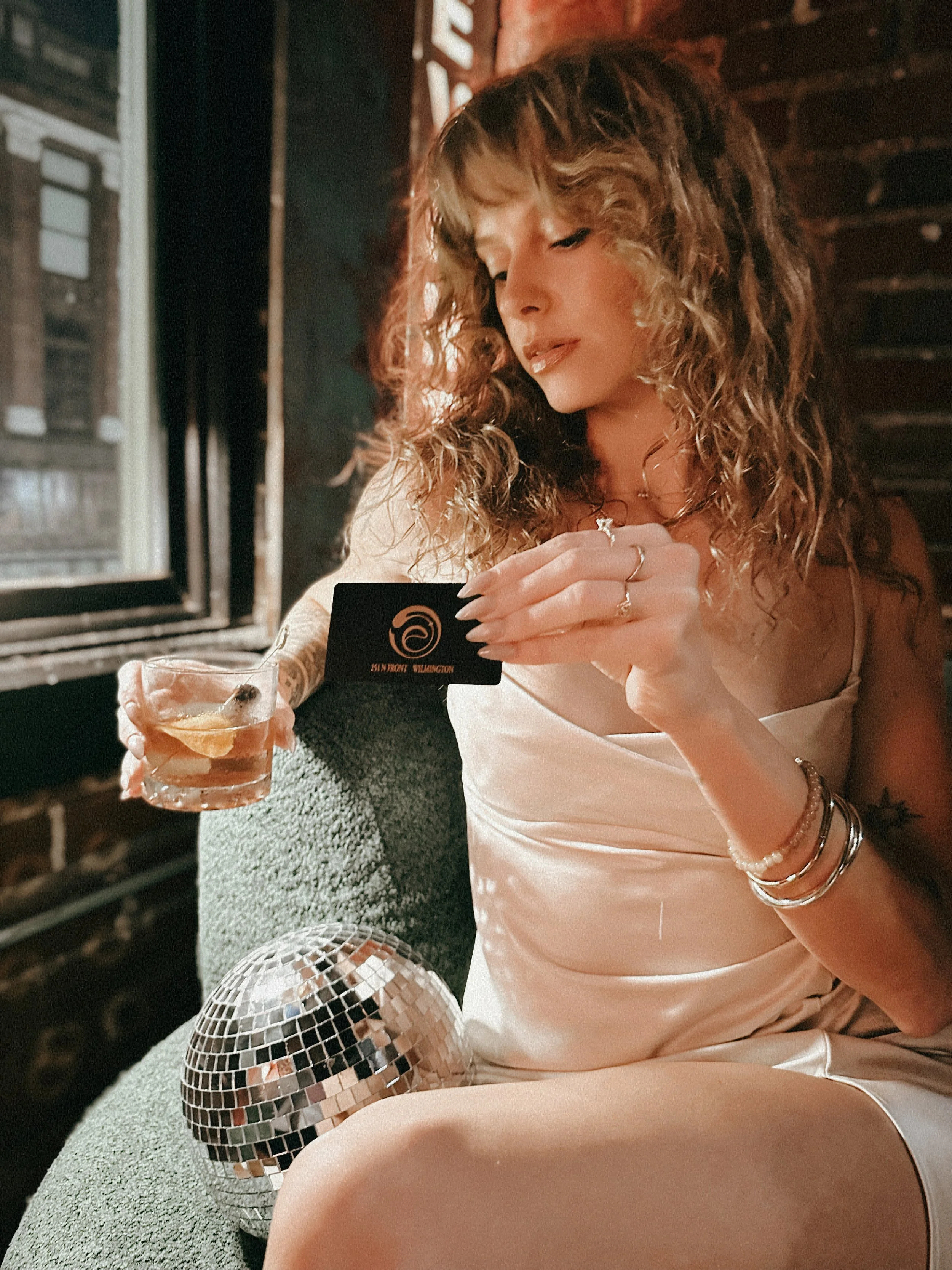 A woman with curly hair sitting on a green chair in a brick-walled room, holding a drink and a black card, with a disco ball on her lap and a window behind her.