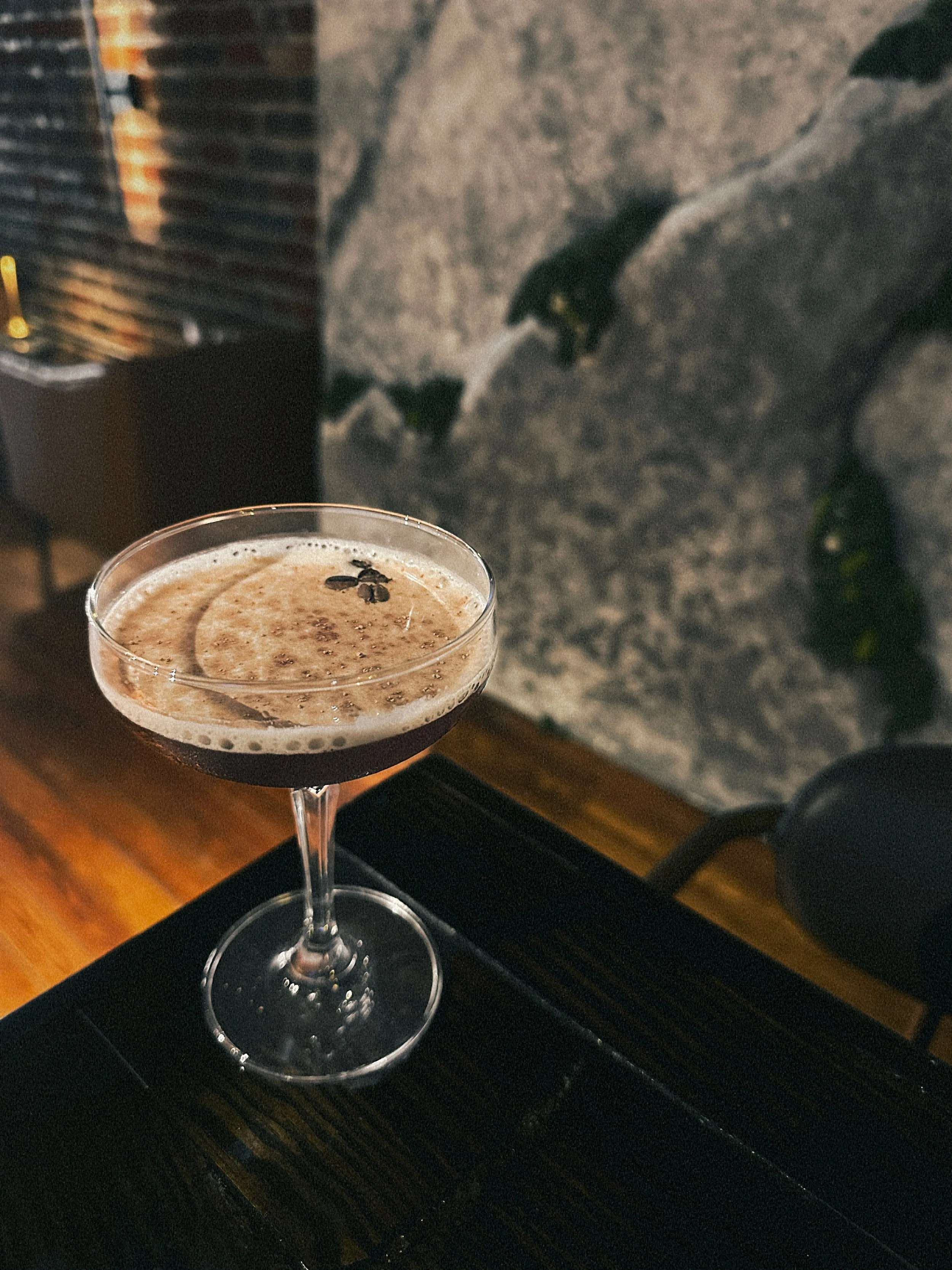 A cocktail in an elegant glass with dark liquid, topped with foam and coffee beans, placed on a black tray.