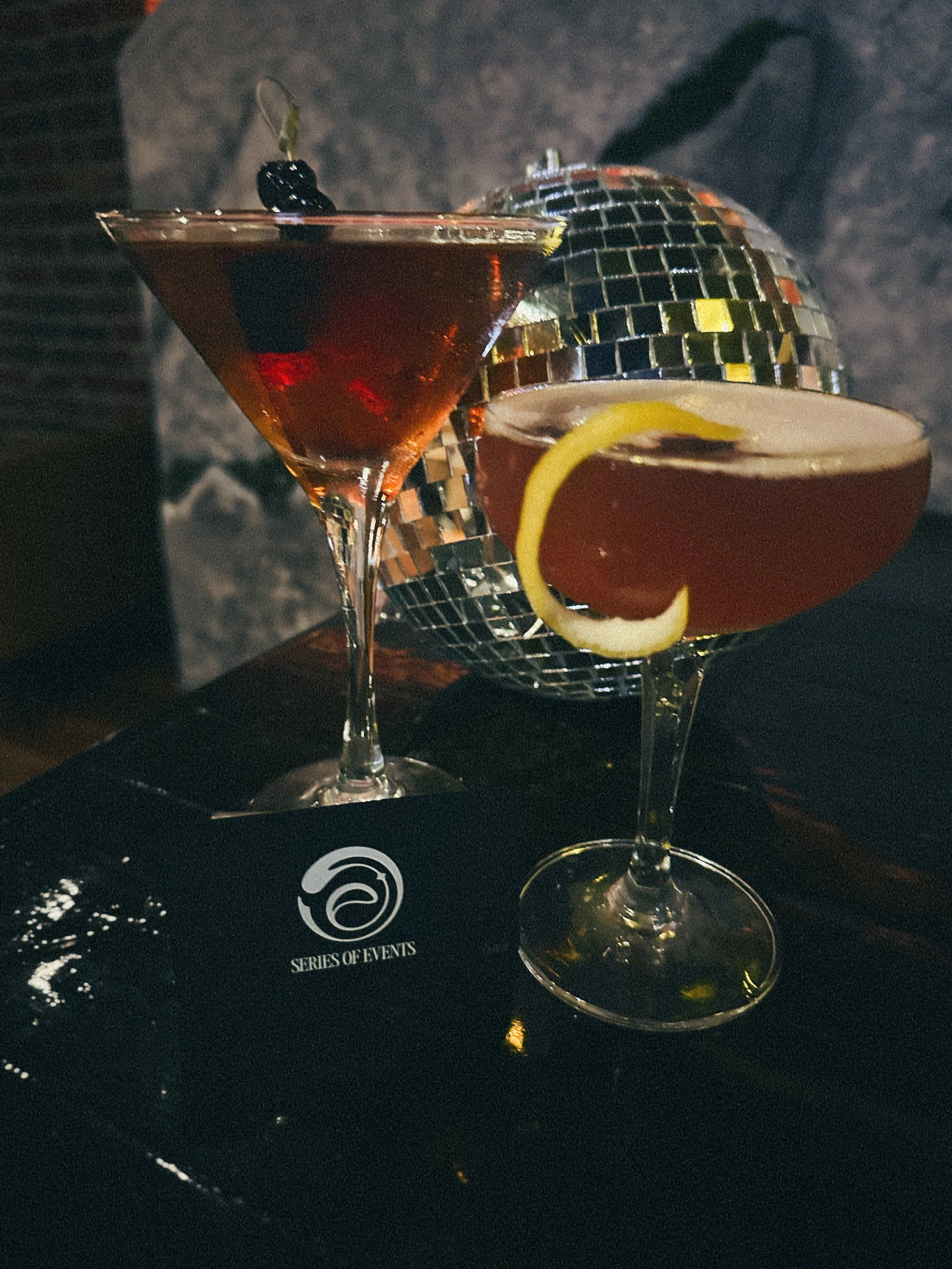 Two cocktails in martini glasses with lemon and cherry garnishes, silver disco ball, and a black napkin with a logo reading 'Series of Events' on a dark surface.