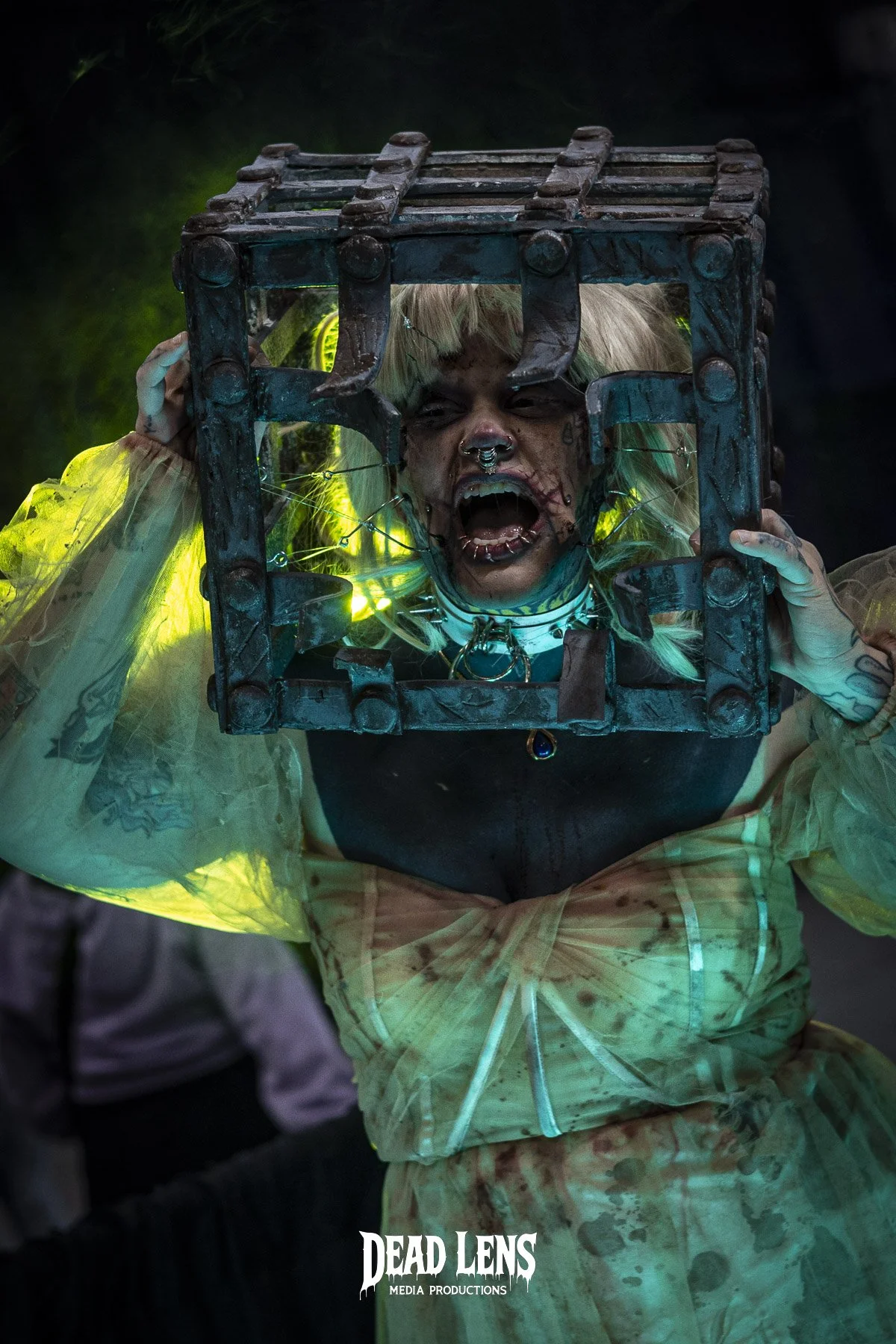 Performer wearing a blonde wig and makeup, holding a metal cage over her head, shouting or screaming during a horror-themed event or performance, with glowing lights and dark background.