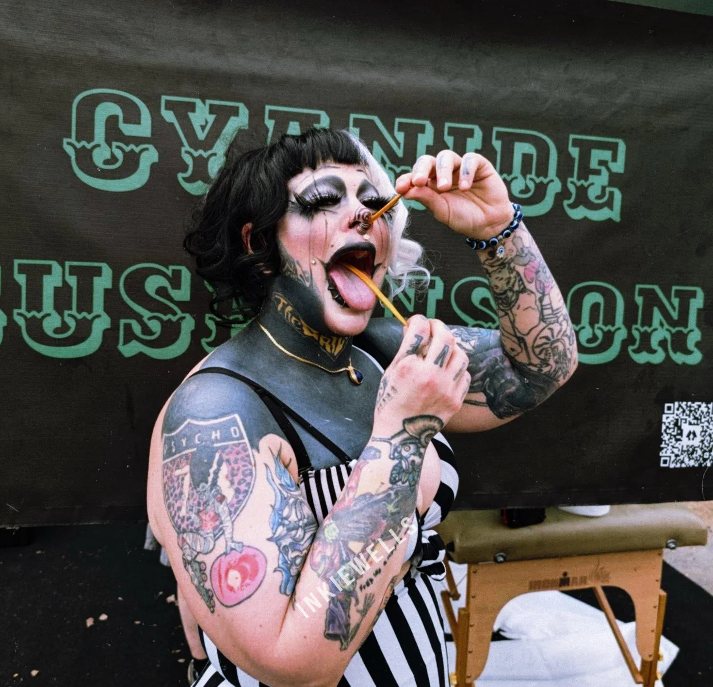 Tattooed woman with makeup and black hair smiling and holding lollipops in her mouth and hand, standing in front of a black background with green text.