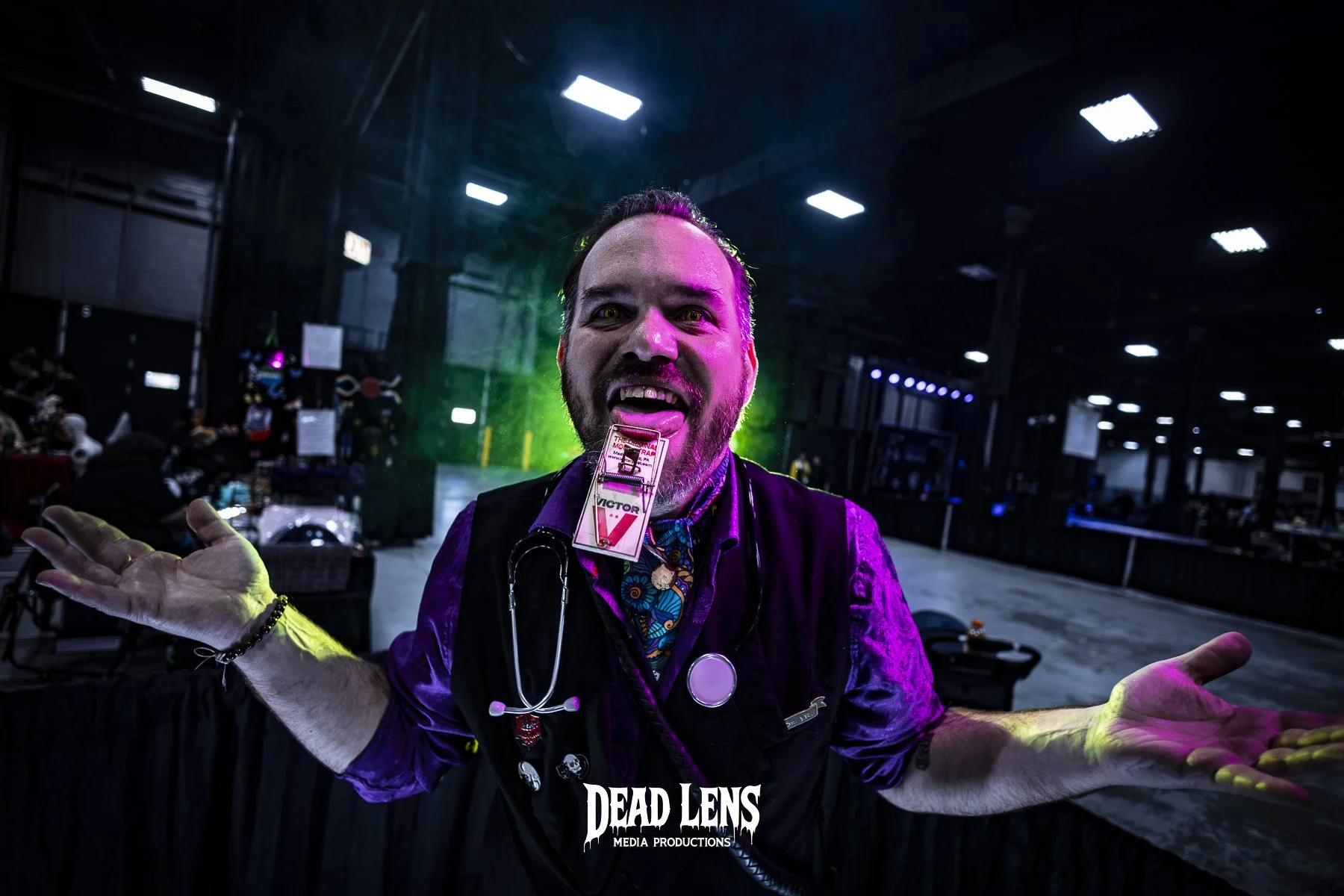 A man with a beard and short dark hair inside a dimly lit convention hall, making a playful face with his mouth open. He wears a stethoscope around his neck, a name badge, and a patterned blue shirt under a purple jacket. The background includes colo