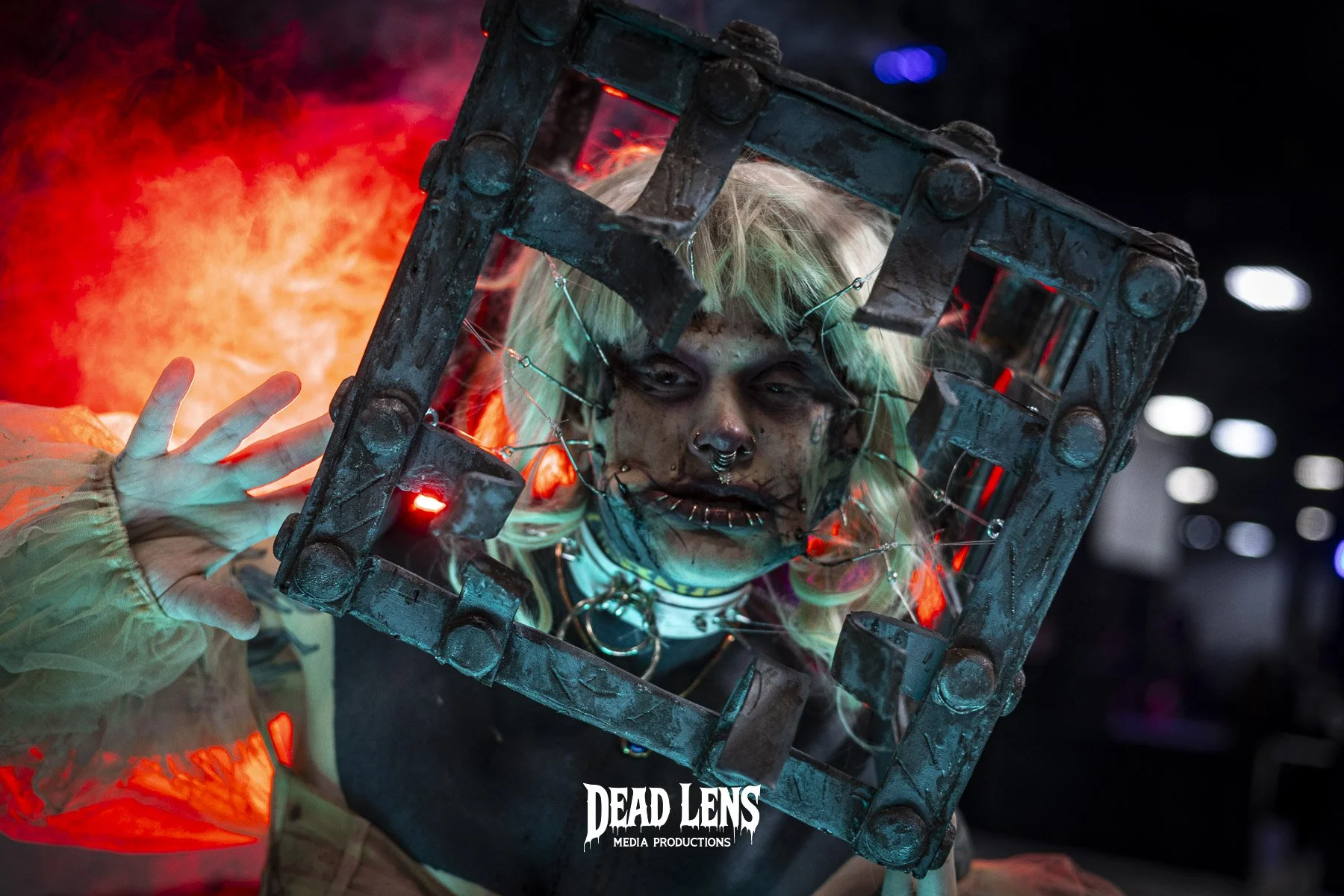 Person with heavy makeup and body modifications holding a metal cage in front of their face, with colorful lighting and a dark background. The person has blonde hair, piercings, and is wearing a choker. The photo has a gritty, intense atmosphere.