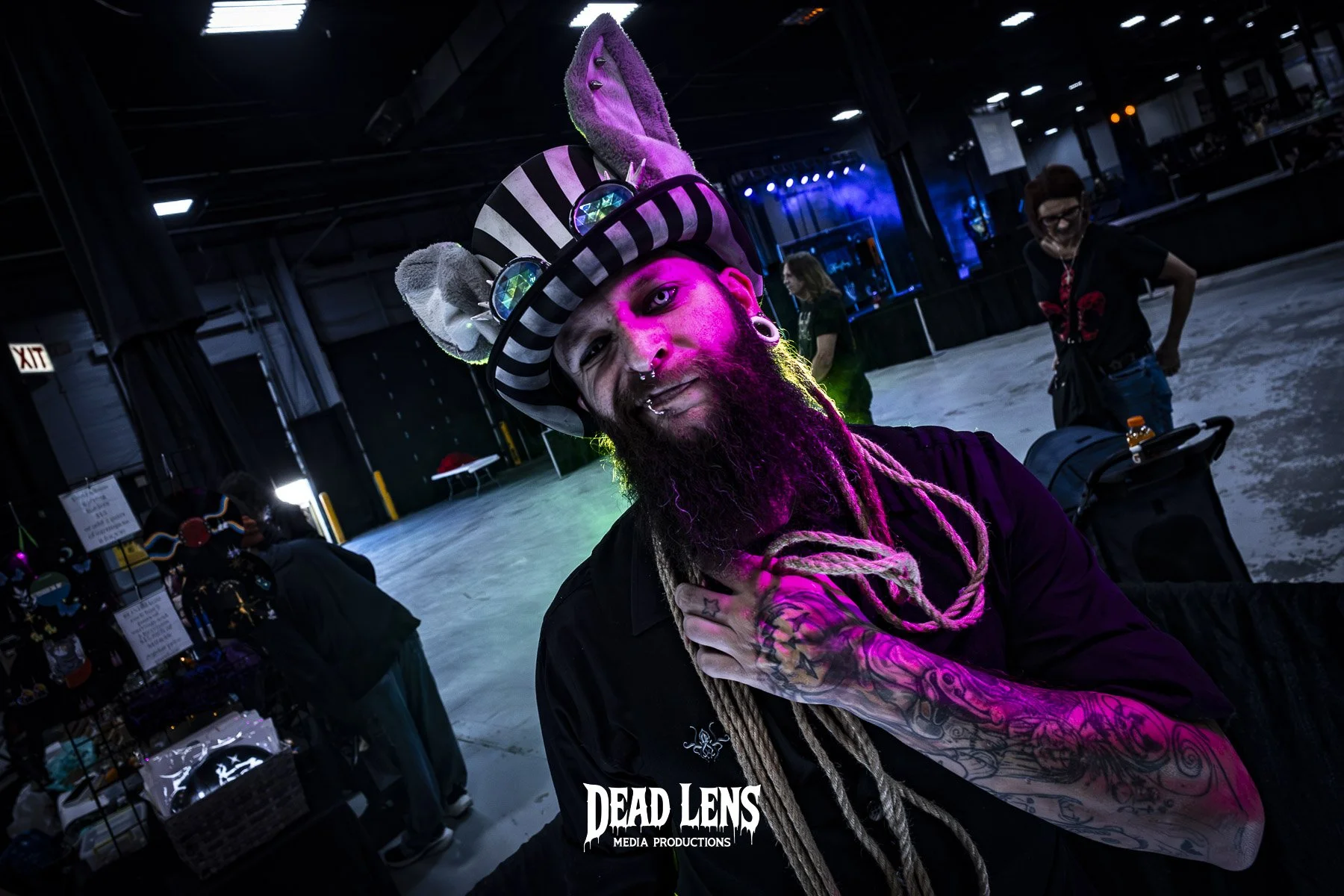 Man with a beard, colorful tattoos, and stretched earlobes, wearing a striped hat with plush bunny ears and goggles, at an indoor event, lit with purple and blue lights.