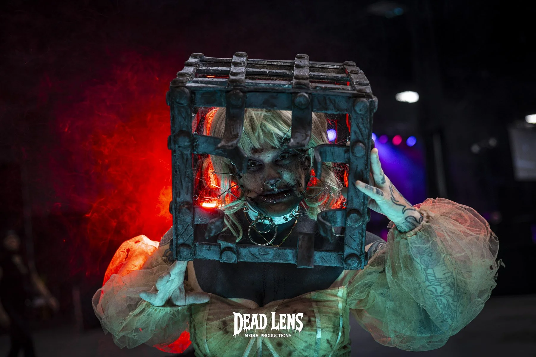 Person with blonde hair and tattoos holding a metal cage in front of their face, with red and purple lights in the background, at a media production event.