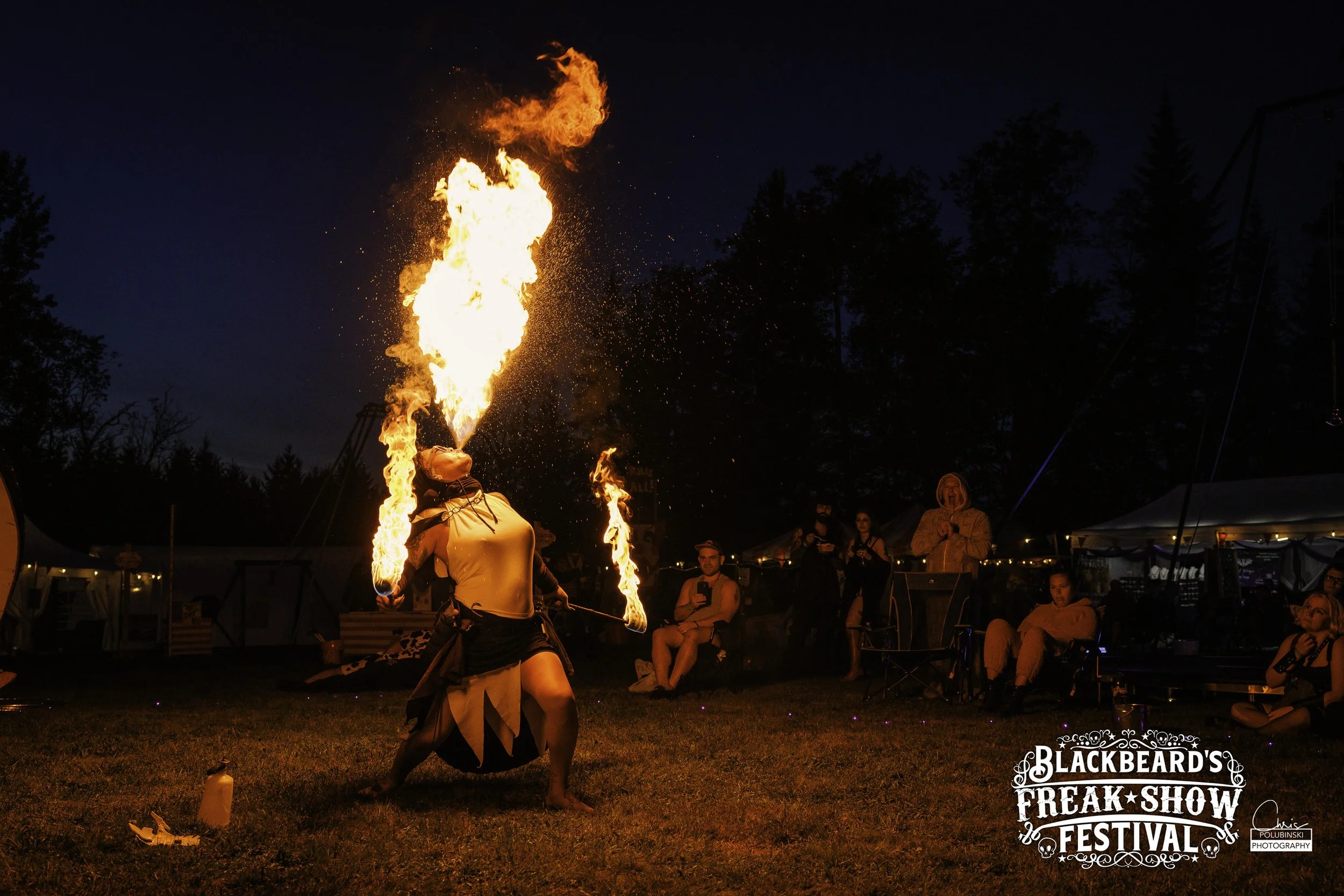 A fire performer at Blackbeard's Freak Show Festival during nighttime, creatively breathing fire with large flames, while a crowd watches in the background near tents and trees.