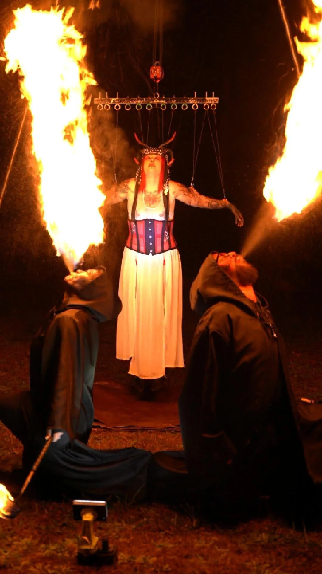 Performer in elaborate costume with horns, suspended in the air, breathing fire as two individuals kneel on the ground with fire torches in their hands.