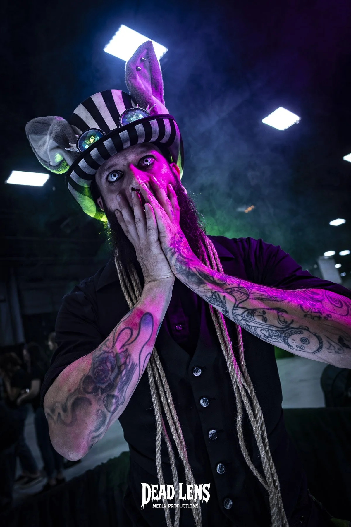 A person wearing a striped top hat with bunny ears and goggles, with long dreadlocks, tattoos on arms, and pale makeup, holding their face with both hands in a surprised or shocked pose under colorful stage lighting.