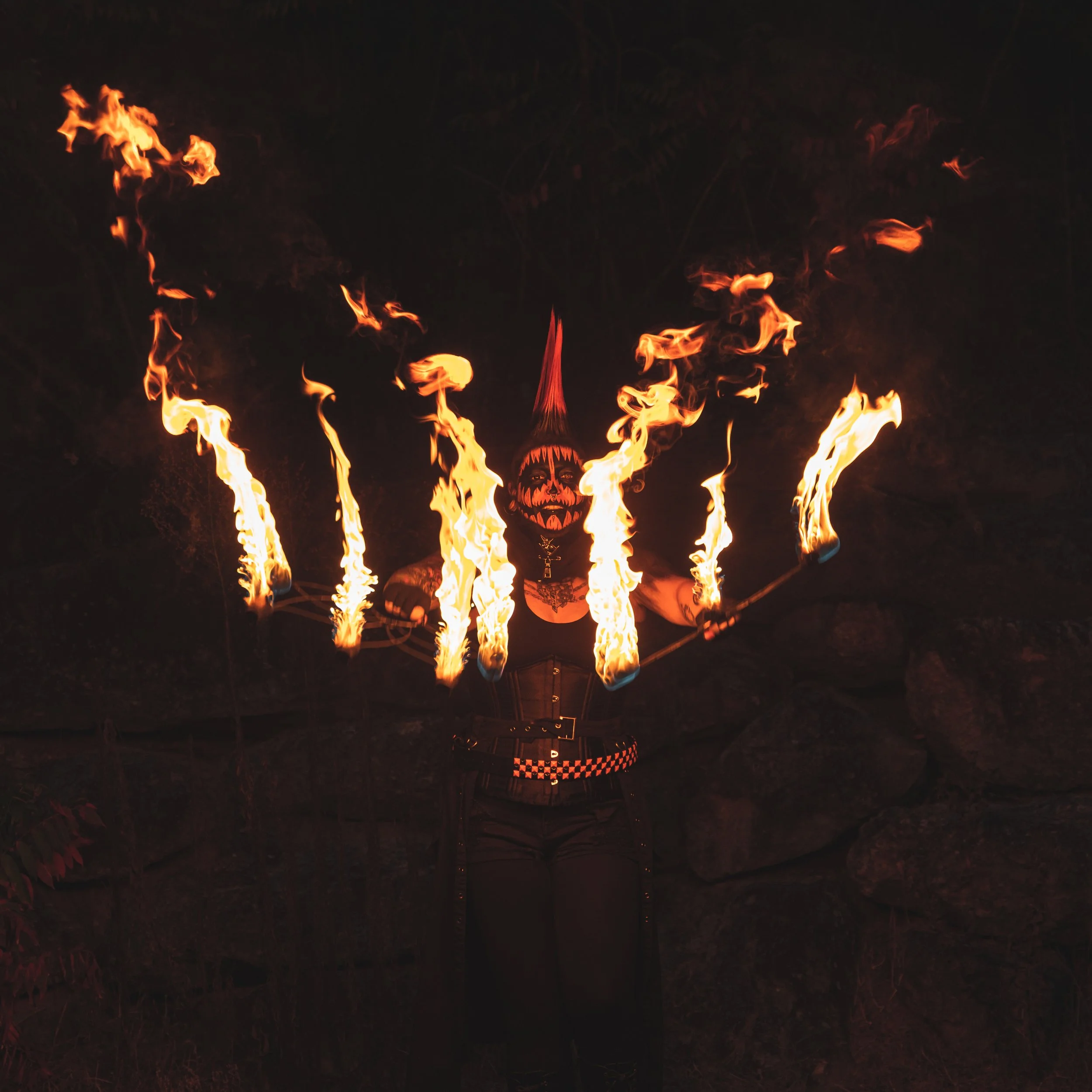 A person dressed in black with face paint resembling a pumpkin, wearing a tall red and black hat, performs a fire spinning act at night, with flames creating arcs in the dark background.