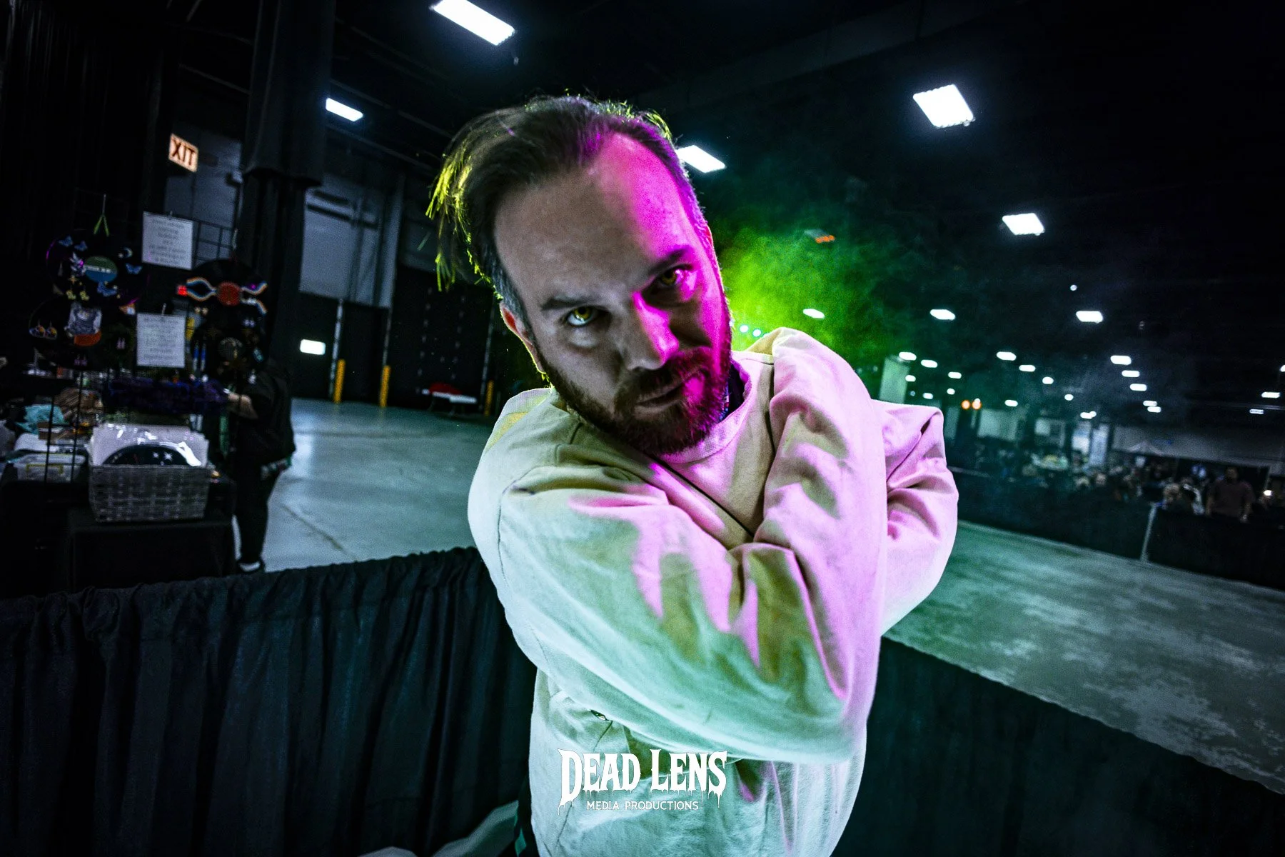 A man with intense eyes and hair styled back, wearing a light-colored hoodie, standing in a dark indoor space with colorful stage lighting, crossing his arms and leaning forward.
