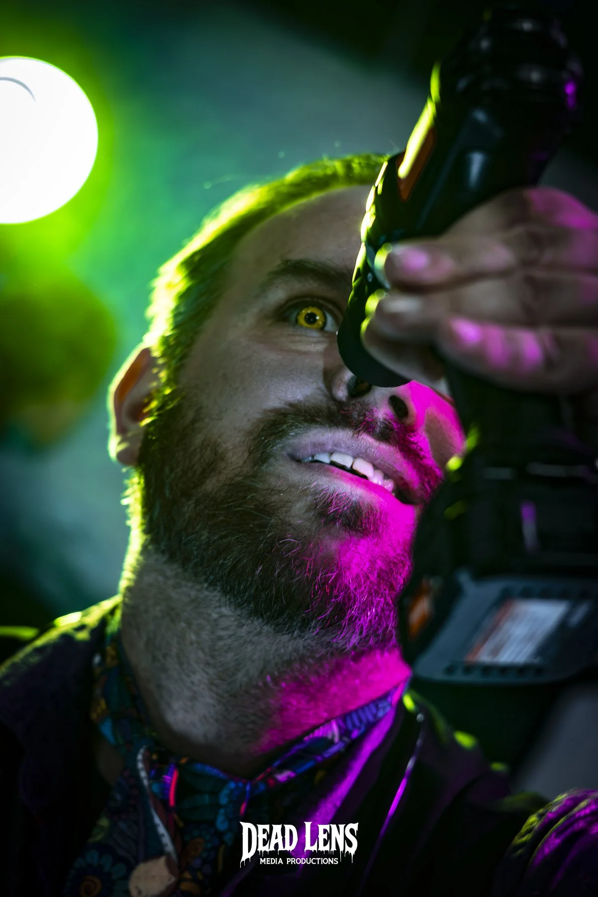 A man with a beard and mustache, holding a camera close to his face, with colorful neon lighting illuminating his features and yellow eye, in a vibrant setting.