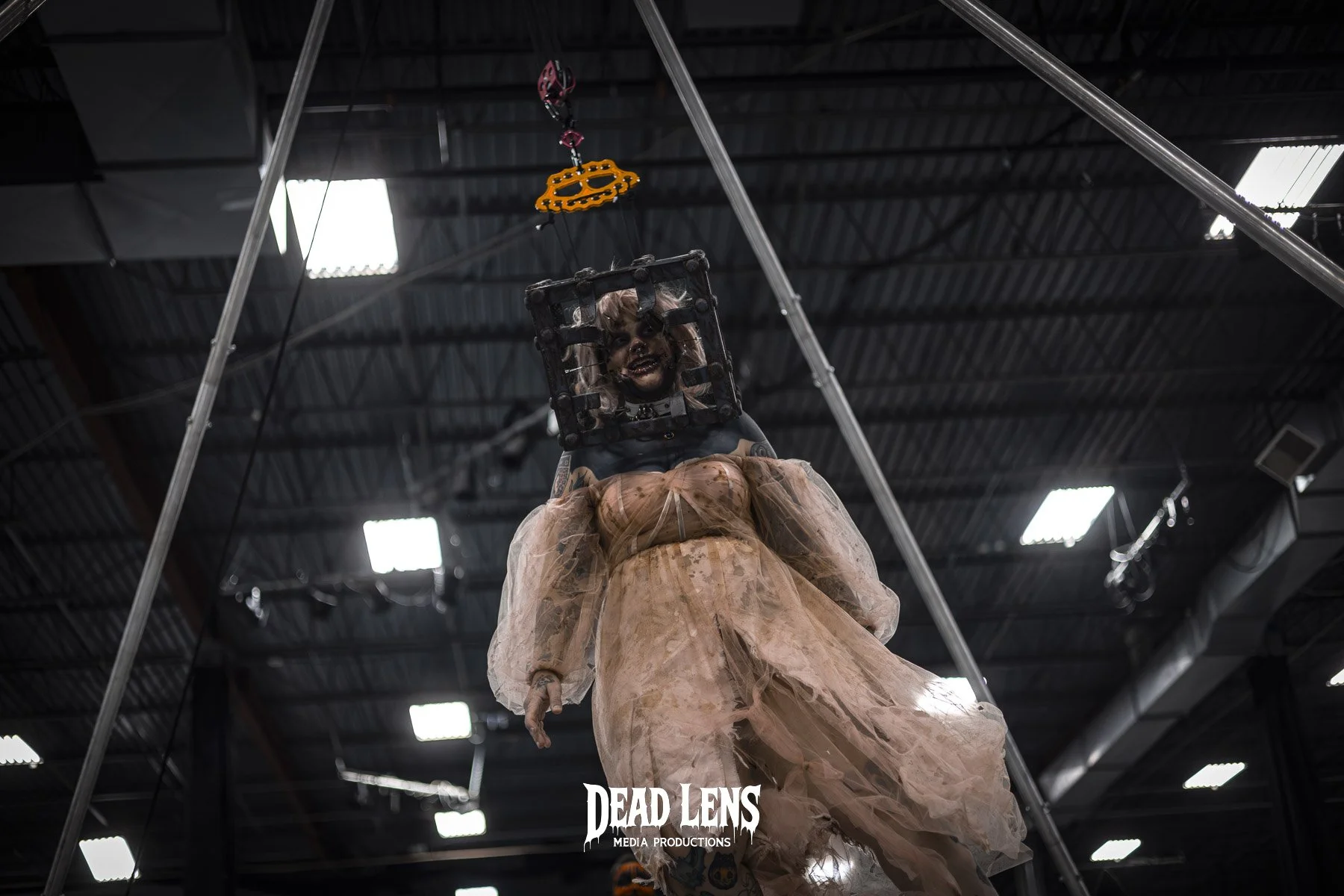 A person dressed as a doll or puppet, hanging inside a metal frame in an industrial-looking space with a dark ceiling and bright lights.