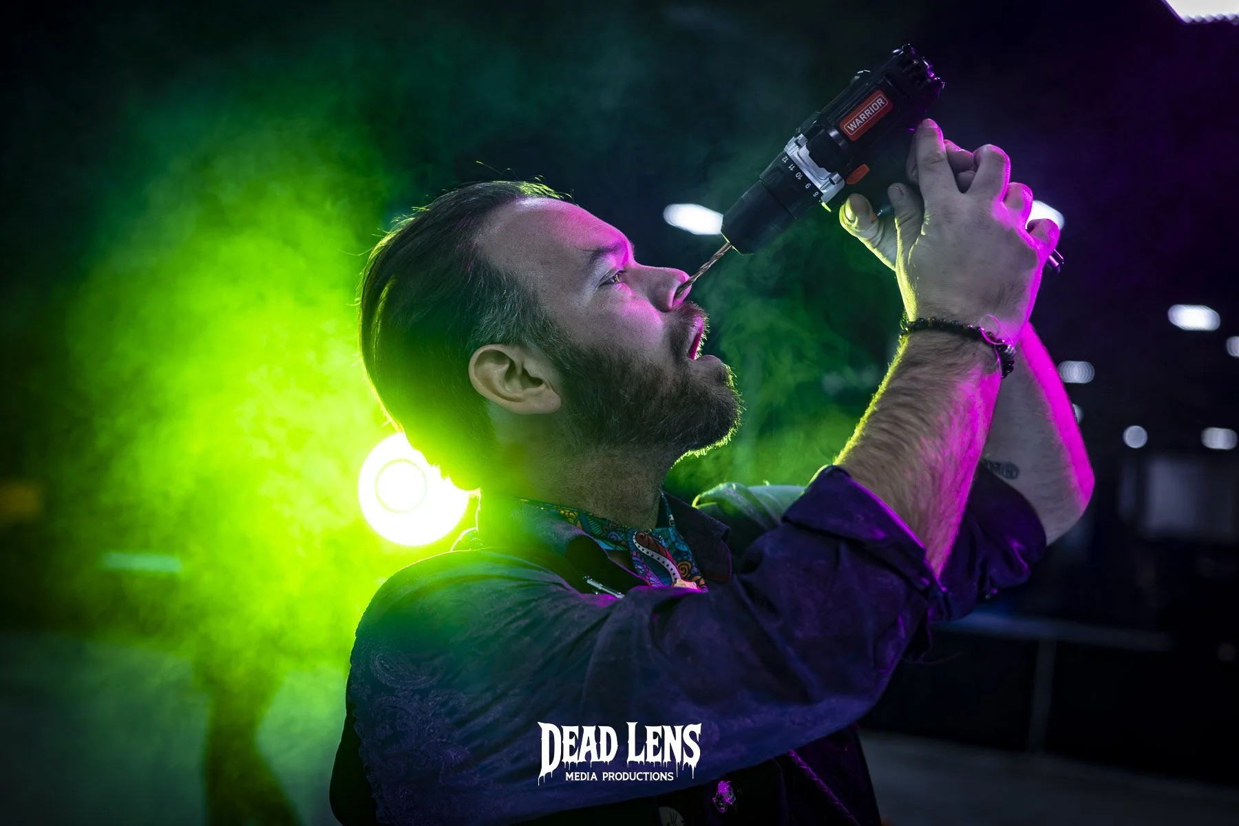 A man with a beard and styled hair is holding a drill-like device to his nose in a colorful setting with green, purple, and pink lighting. The background is dark with some lights. The text "Dead Lens Media Productions" is at the bottom.