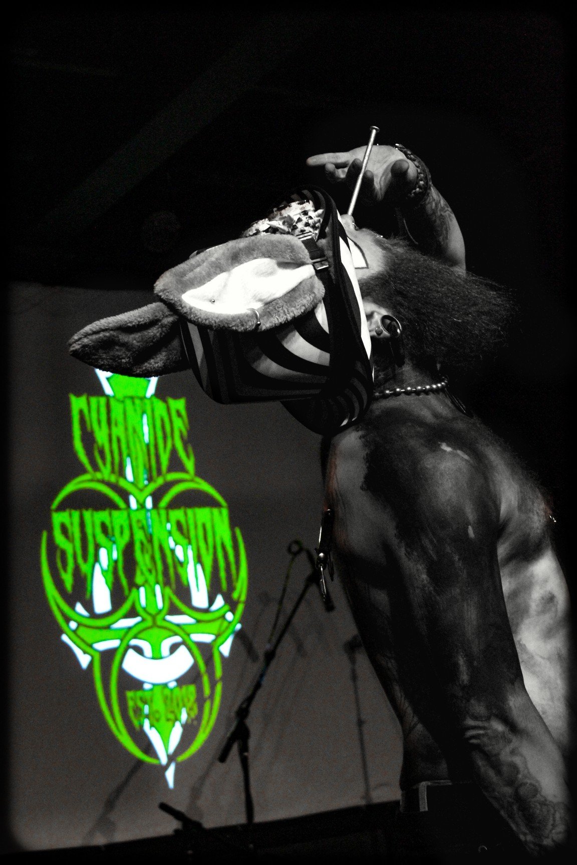 A person with tattoos and dark hair, wearing jewelry, holds a stuffed animal and a cigarette, with a neon green sign in the background that says 'CHANGE' and 'SUSPENSION'.