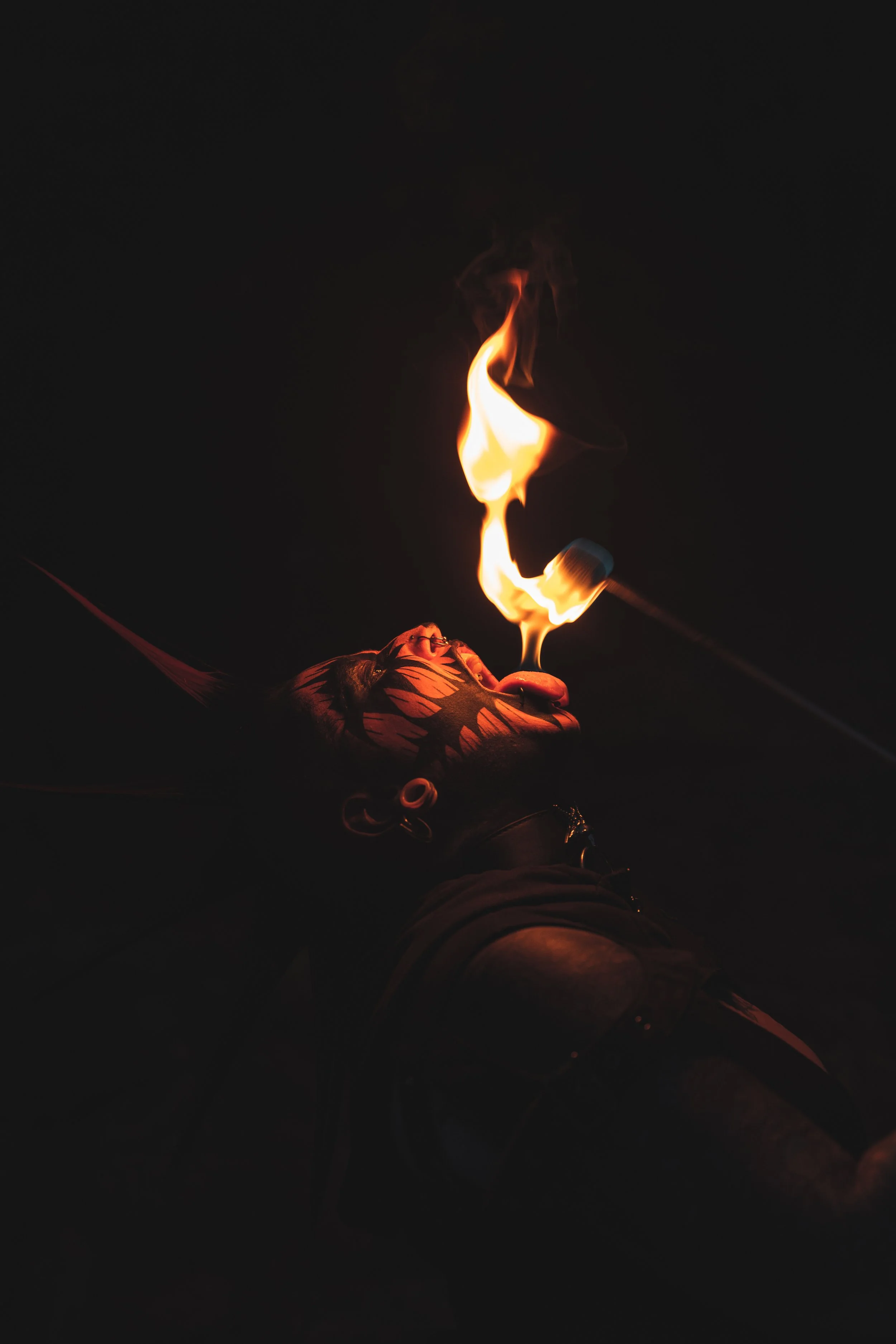 A person lying down with face painted, wearing earrings, and holding a lit matchstick in the mouth, producing a large flame.