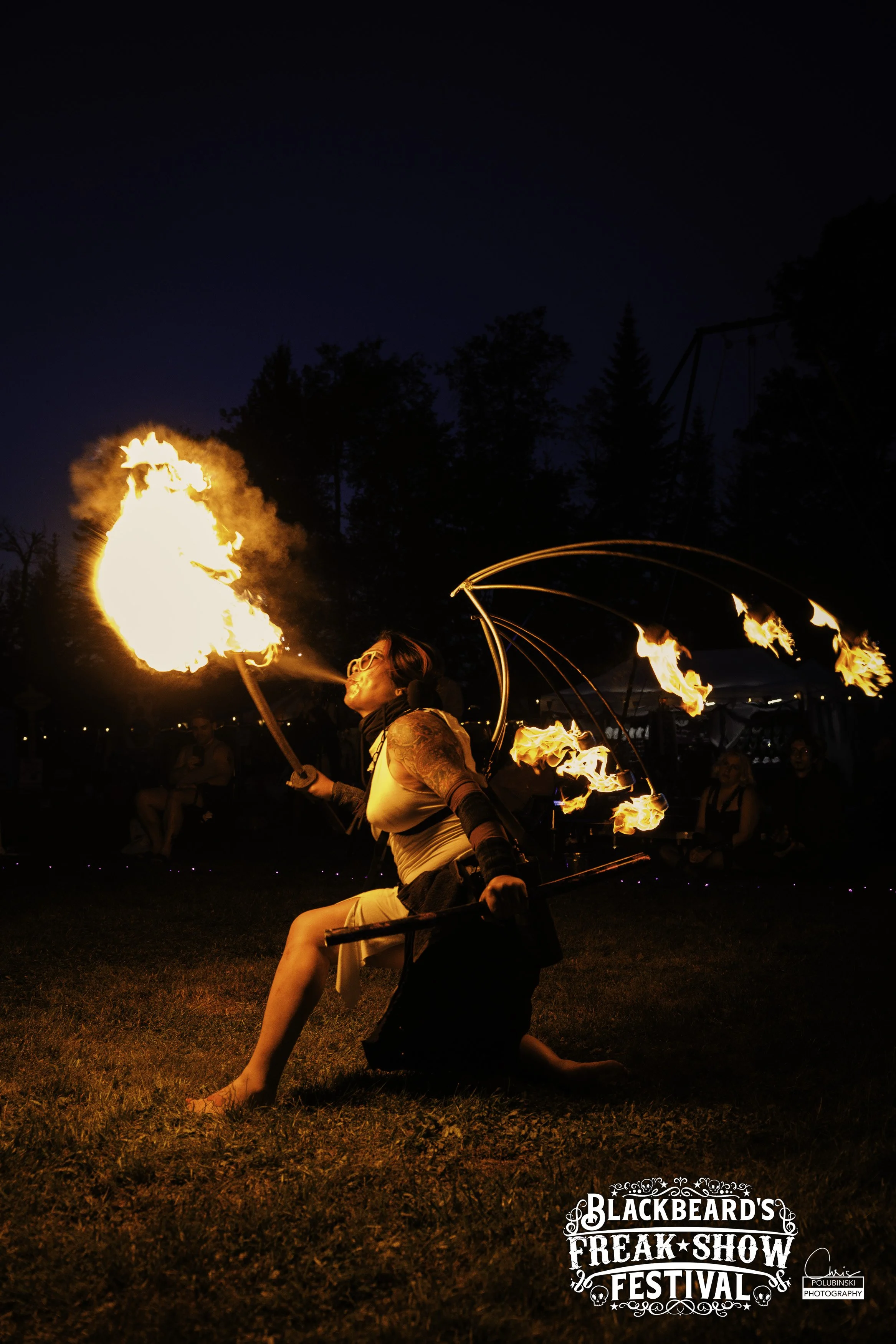 A female fire performer in a black dress and arm coverings burning fire, performing at night during the Blackbeard's Freak Show Festival, with audience members in the background.