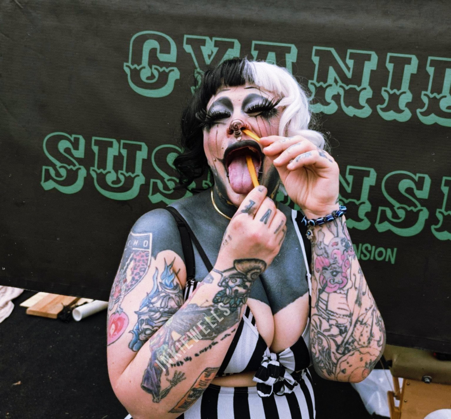 A heavily tattooed person with dramatic makeup, tongue out, holding candy, and sticking a yellow candy stick into their nose. They are in front of a black and green background with text.