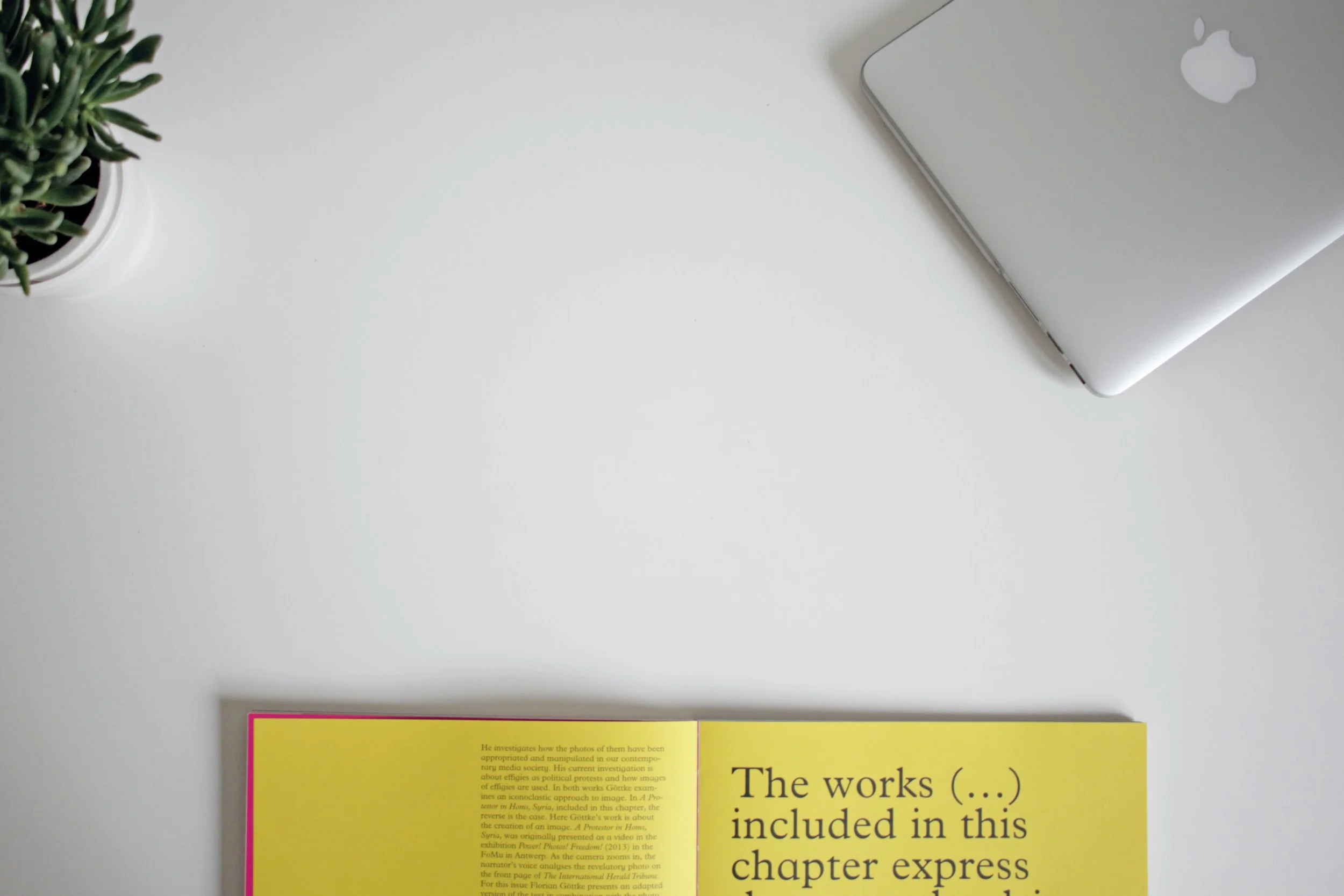 A white desk with a small potted plant on the left, a closed silver MacBook on the right, and an open yellow book with black text at the bottom center.