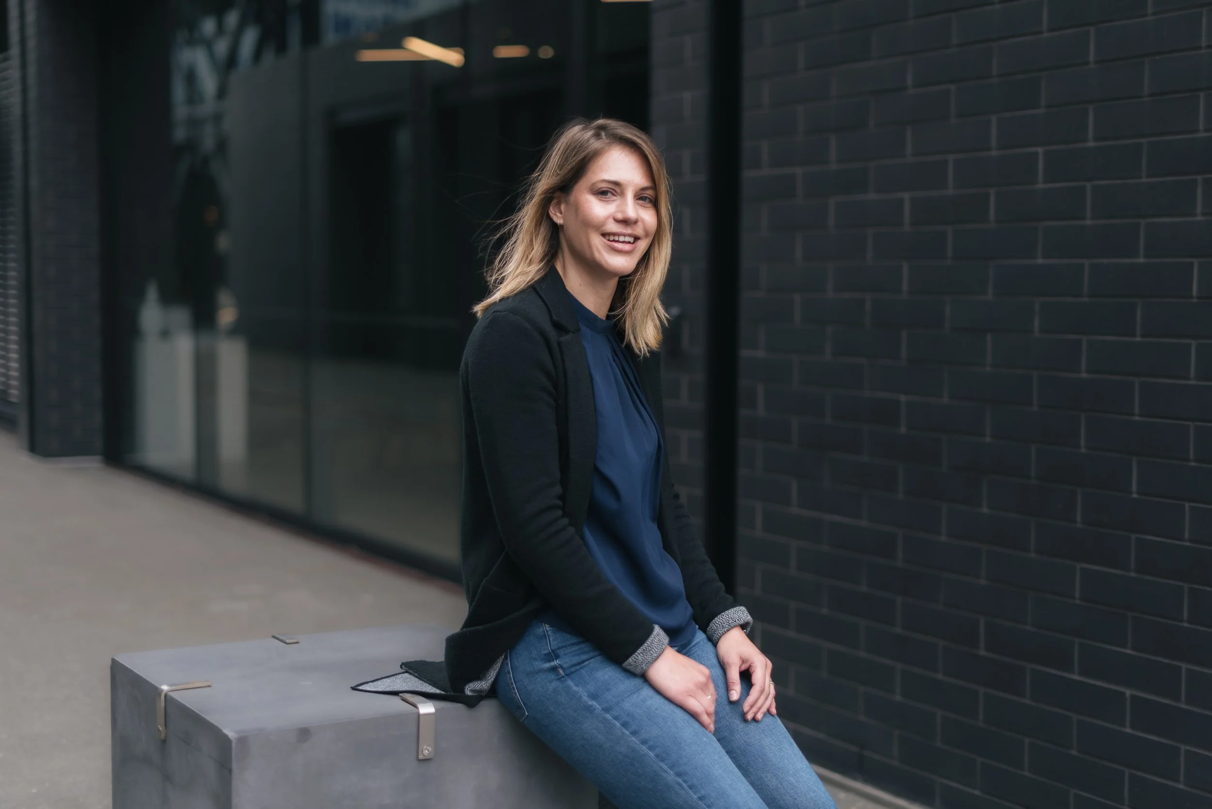A young woman with blonde hair and a black jacket sitting on a gray concrete bench outside a modern building with black brick walls, smiling at the camera.