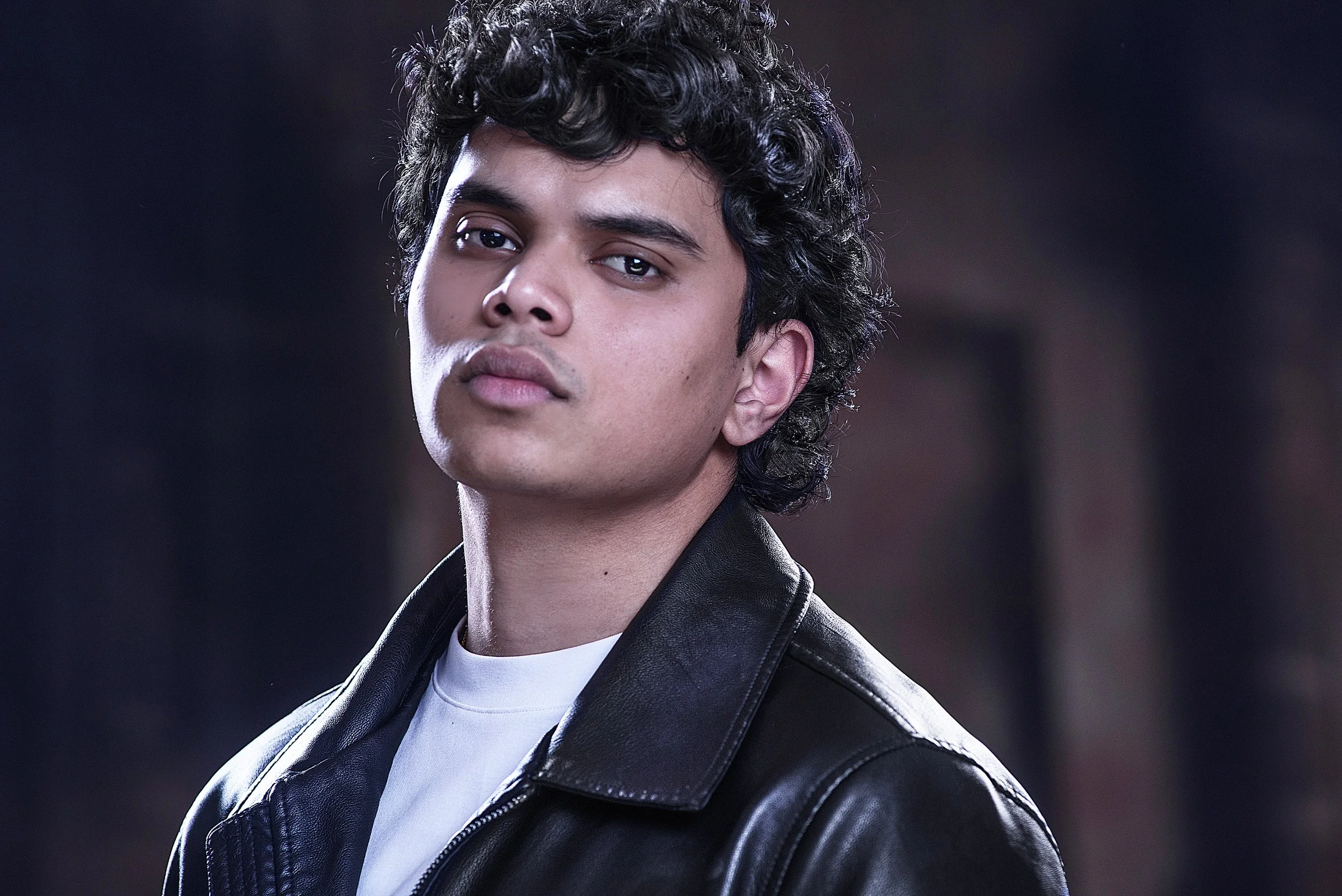 A young man with curly black hair wearing a black leather jacket and a white shirt, looking confidently at the camera against a dark blurred background.